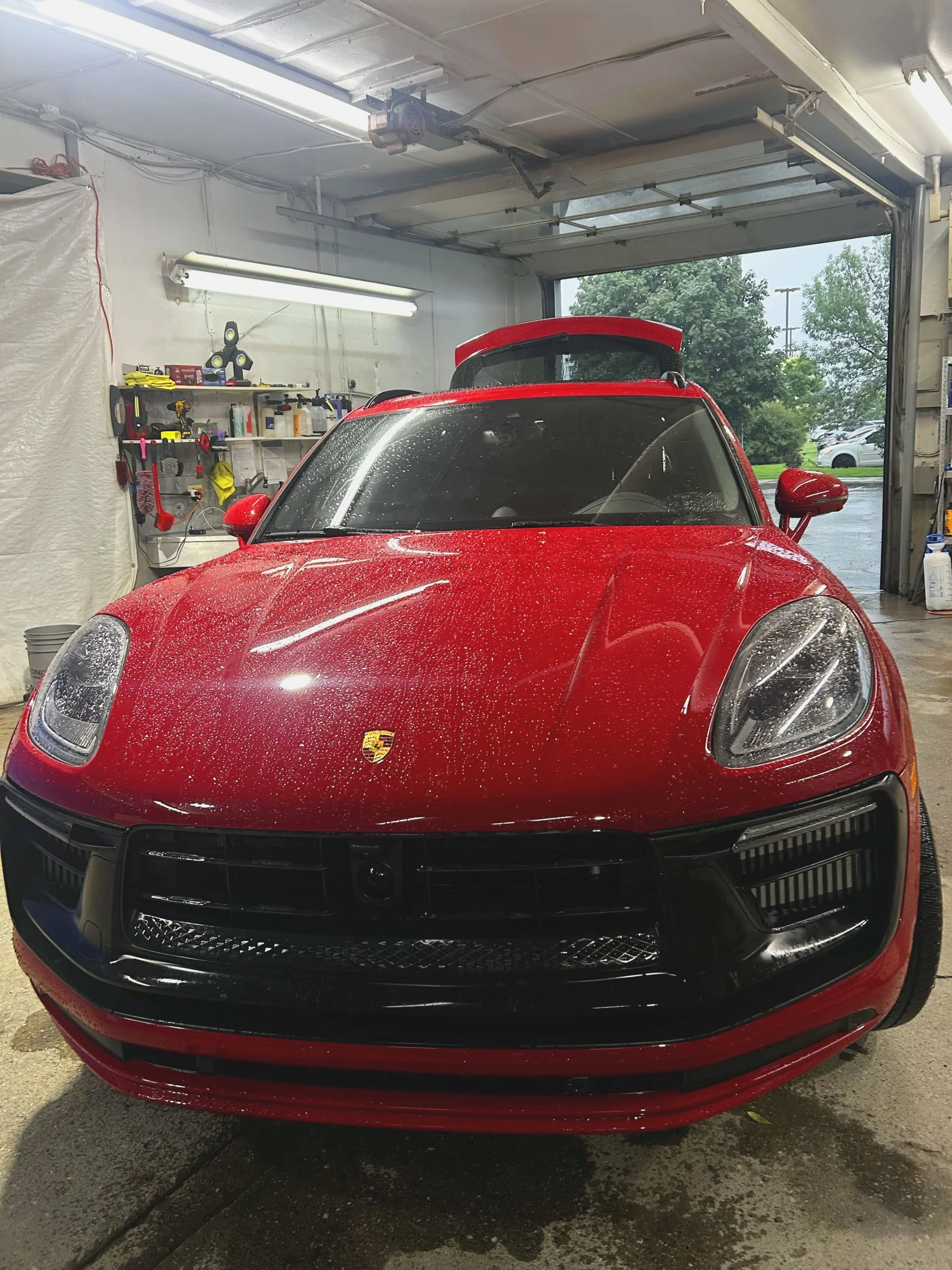 Red sports car with water droplets, parked in a garage with tools and supplies on shelves and an open garage door revealing a parking lot.