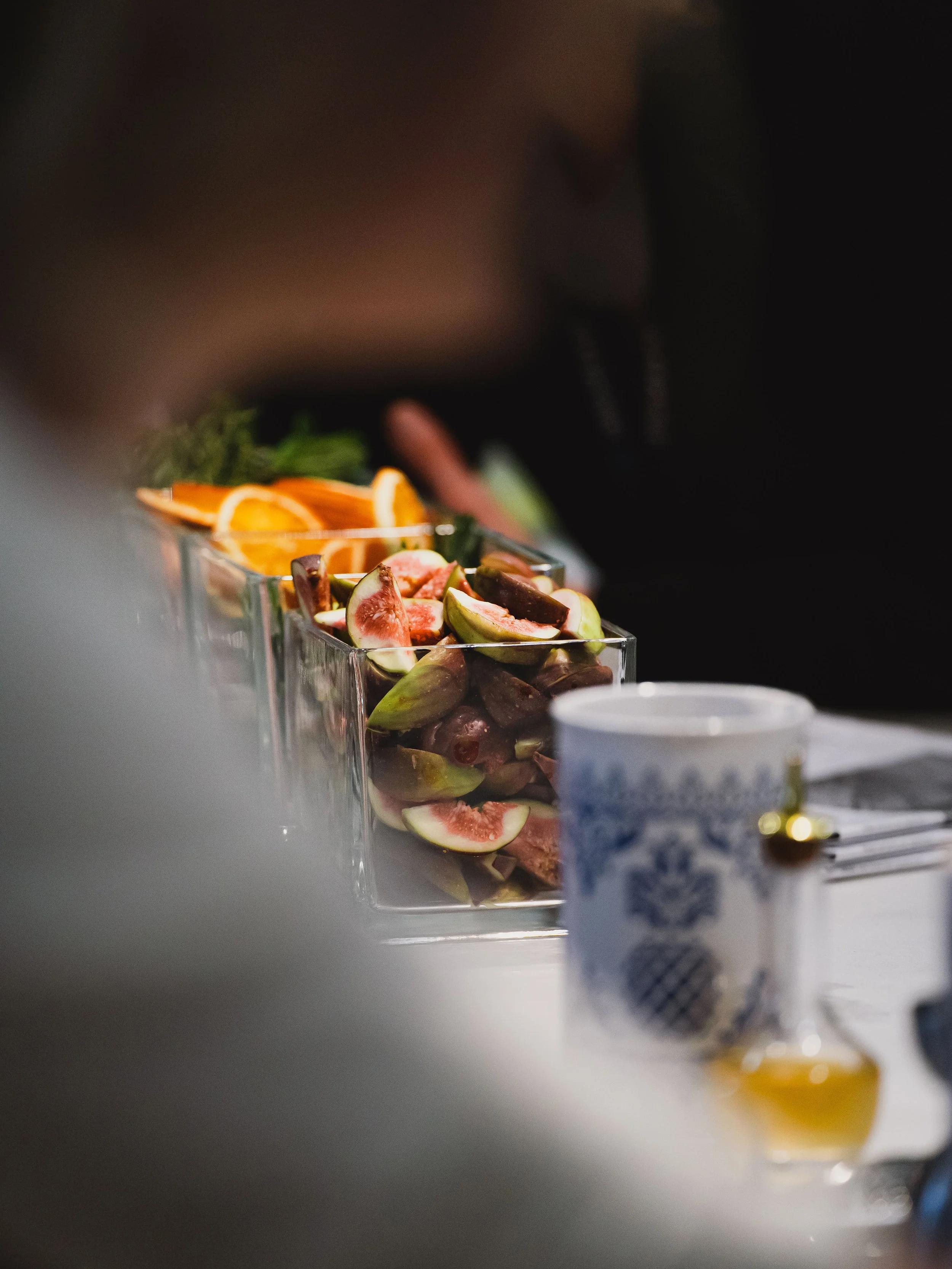 Glass bowls filled with sliced figs and oranges on a table, with a blurry person in the background and a white cup with blue patterns in the foreground.