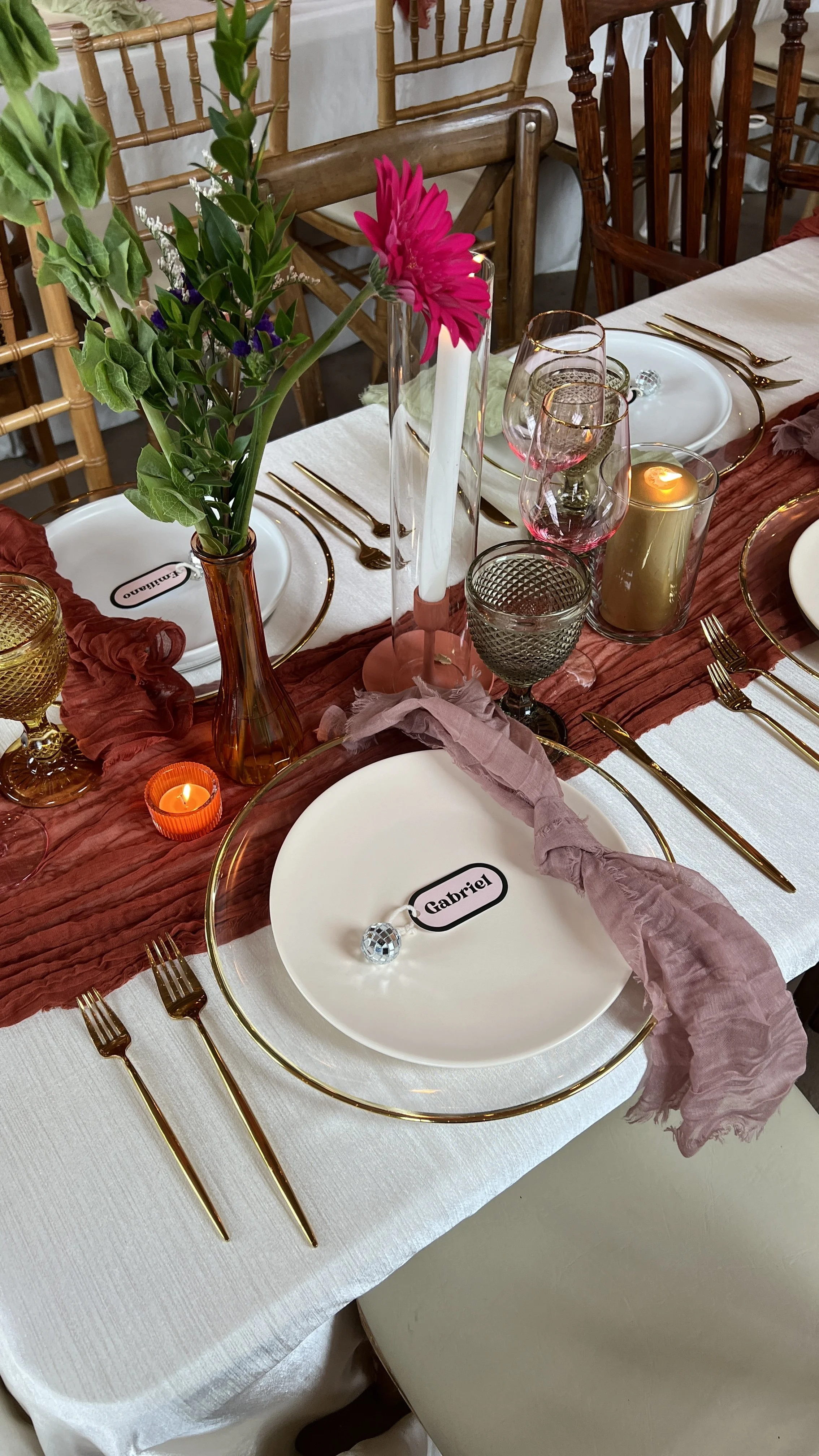 A decorated dining table set with white dinner plates, gold cutlery, and various glasses. There are pink and green floral centerpieces, pink candles, a pink cloth napkin, and a name tag that reads 'Gabriel'.