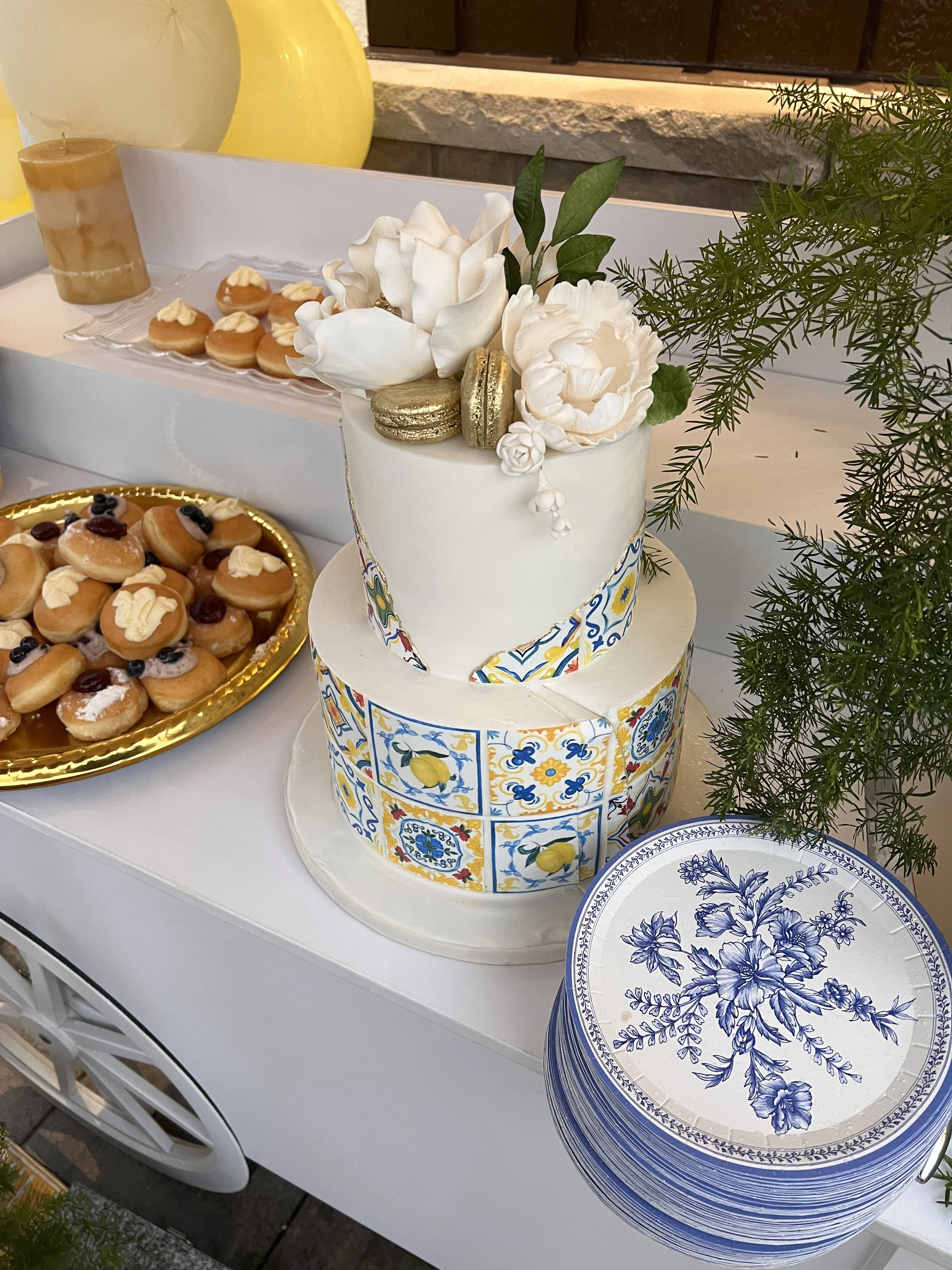 A two-tiered cake decorated with white flowers, gold macarons, and colorful tile-like patterns, surrounded by plates of cookies and a floral-patterned cake stand.