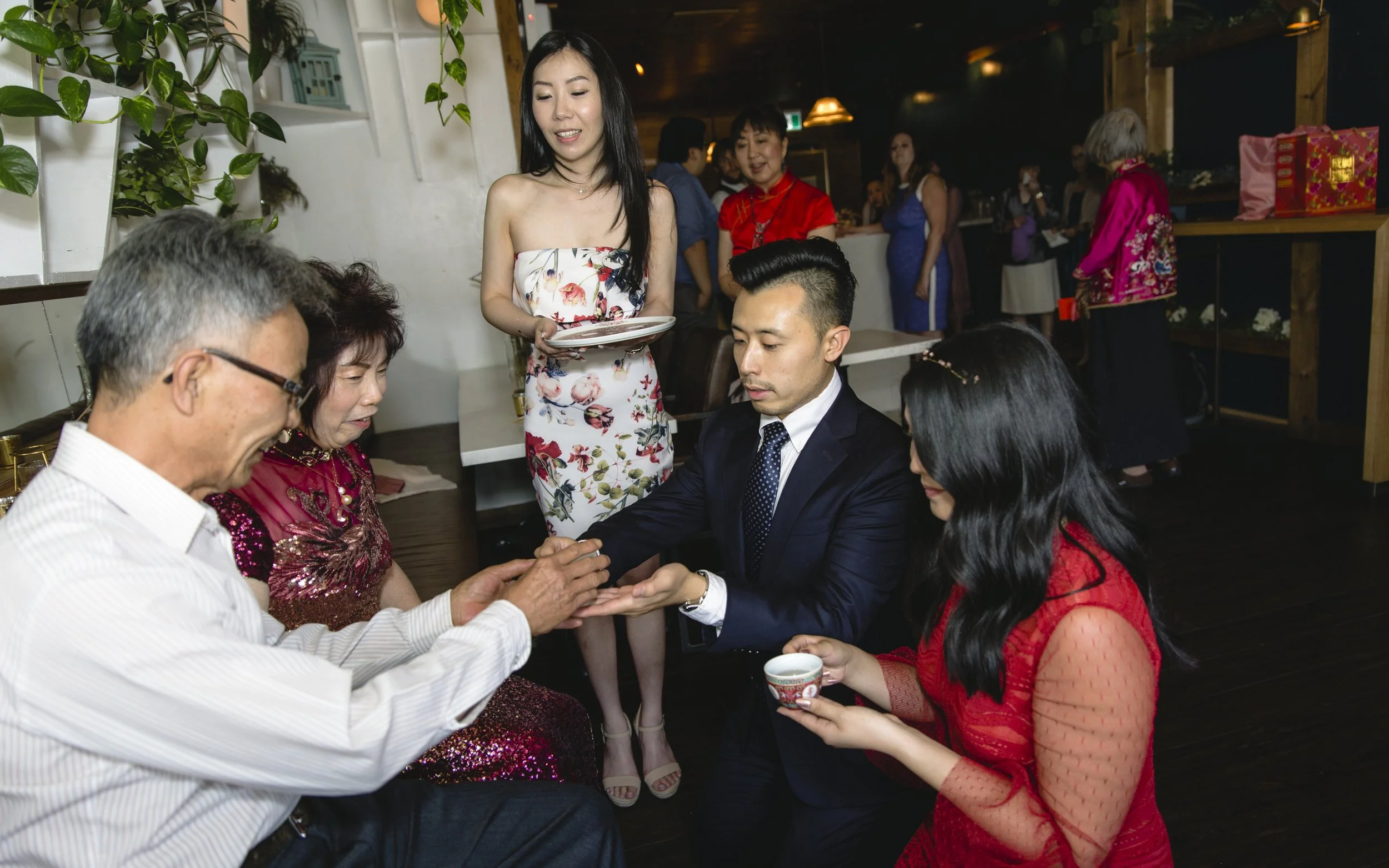 A Korean couple is performing a traditional tea ceremony for their elders during a celebration at a restaurant, with a waitress serving food in the background.