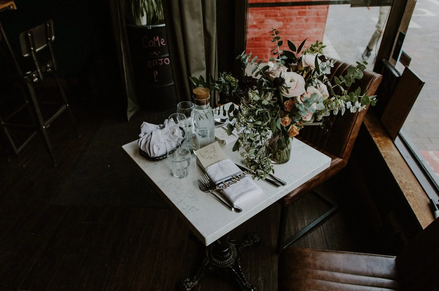 A restaurant table decorated with a large floral arrangement, tableware, and glasses, set near a window with natural light.