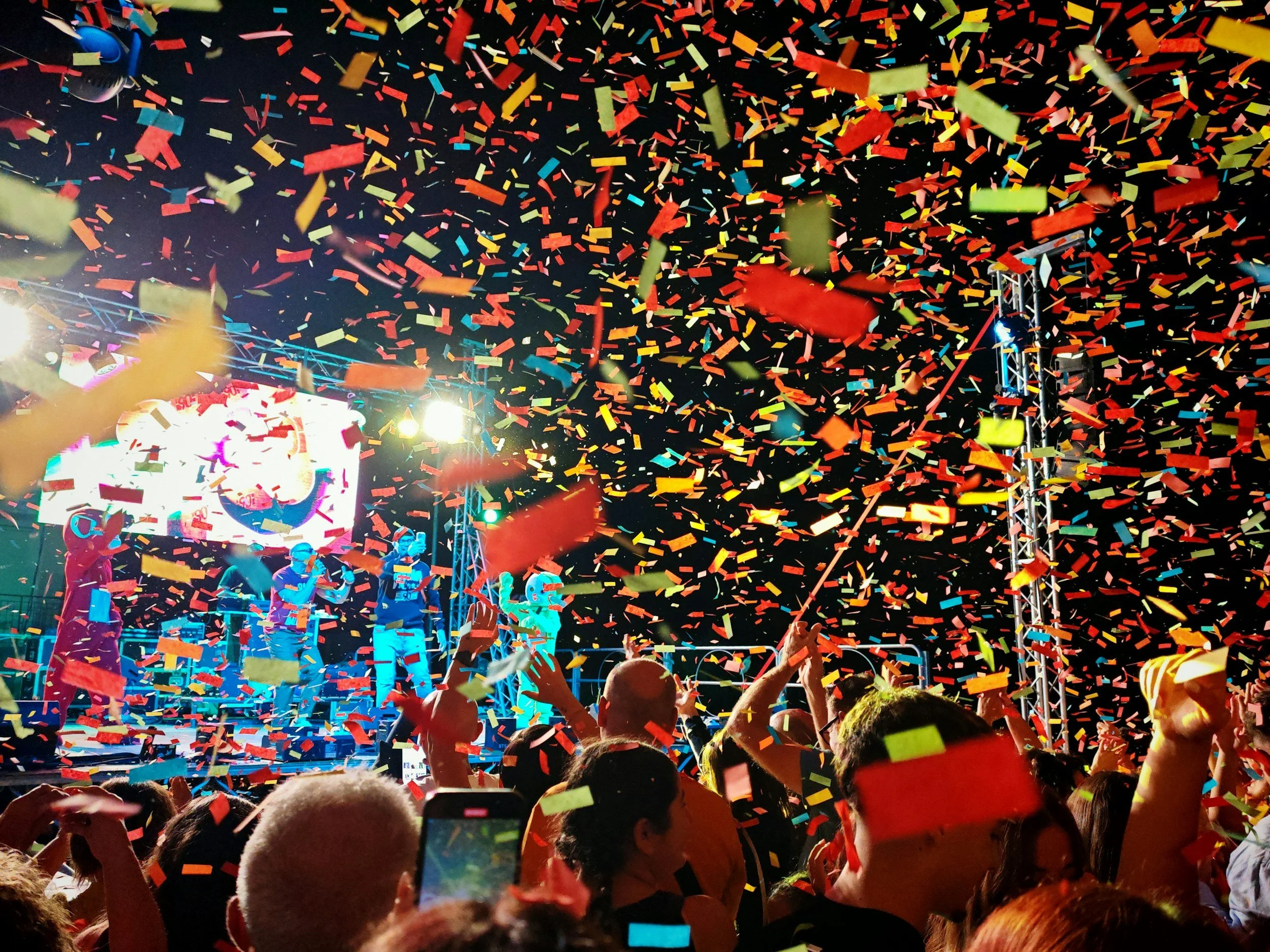 Crowd at a celebration or concert with colorful confetti falling from the sky, stage lights, and performers on stage.