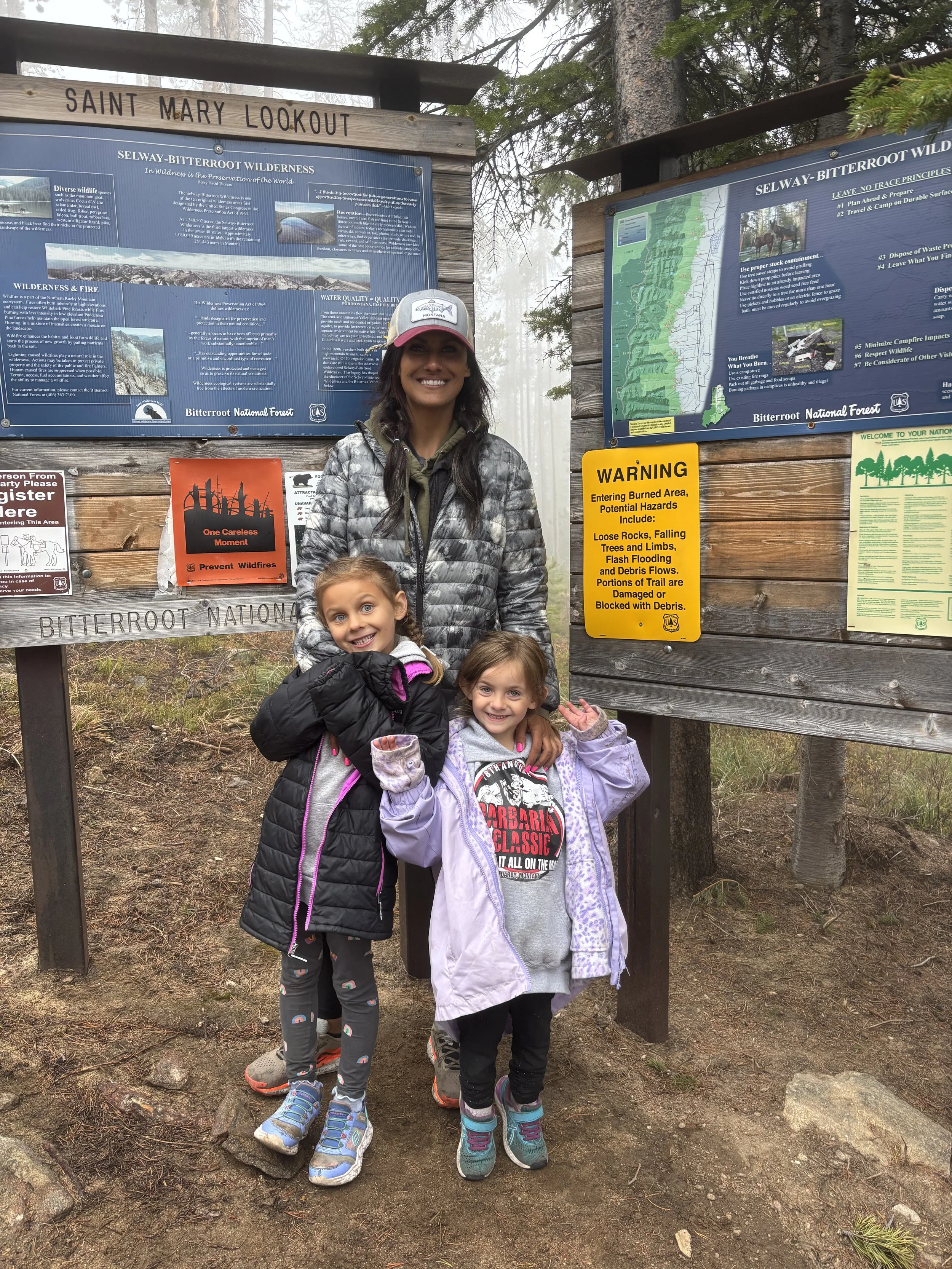 A smiling woman and two young girls posing in front of informational signs at Saint Mary Lookout in Bitterroot National Forest. The area is outdoors, surrounded by trees and fog.