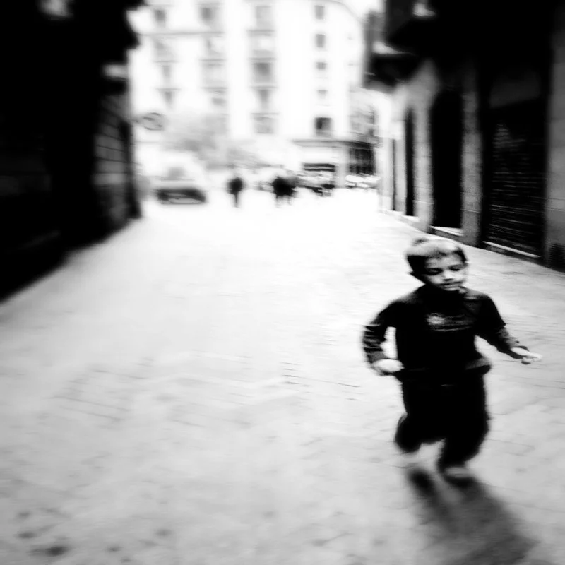 Das Bild zeigt eine unscharfe, schwarz-weiße Aufnahme einer Straßenszene mit einem Jungen im Vordergrund, der auf die Kamera zuläuft, während im Hintergrund Gebäude und andere Personen sichtbar sind.