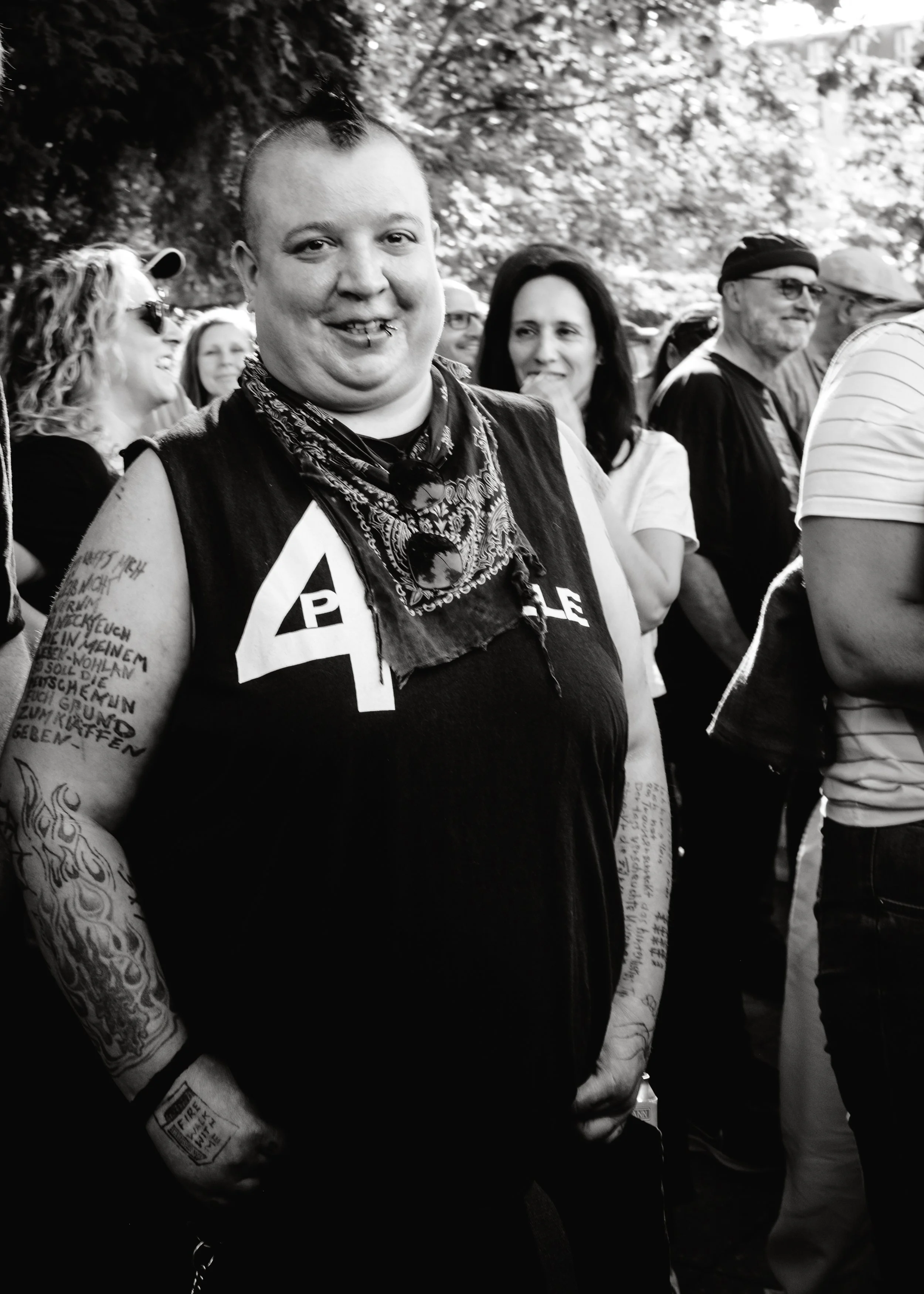 Schwarz-Weiß-Foto einer Person mit Tattoo-Armen, die auf einem Publikumsausflug steht, inmitten von lachenden und entspannten Menschen bei einer Veranstaltung im Freien.