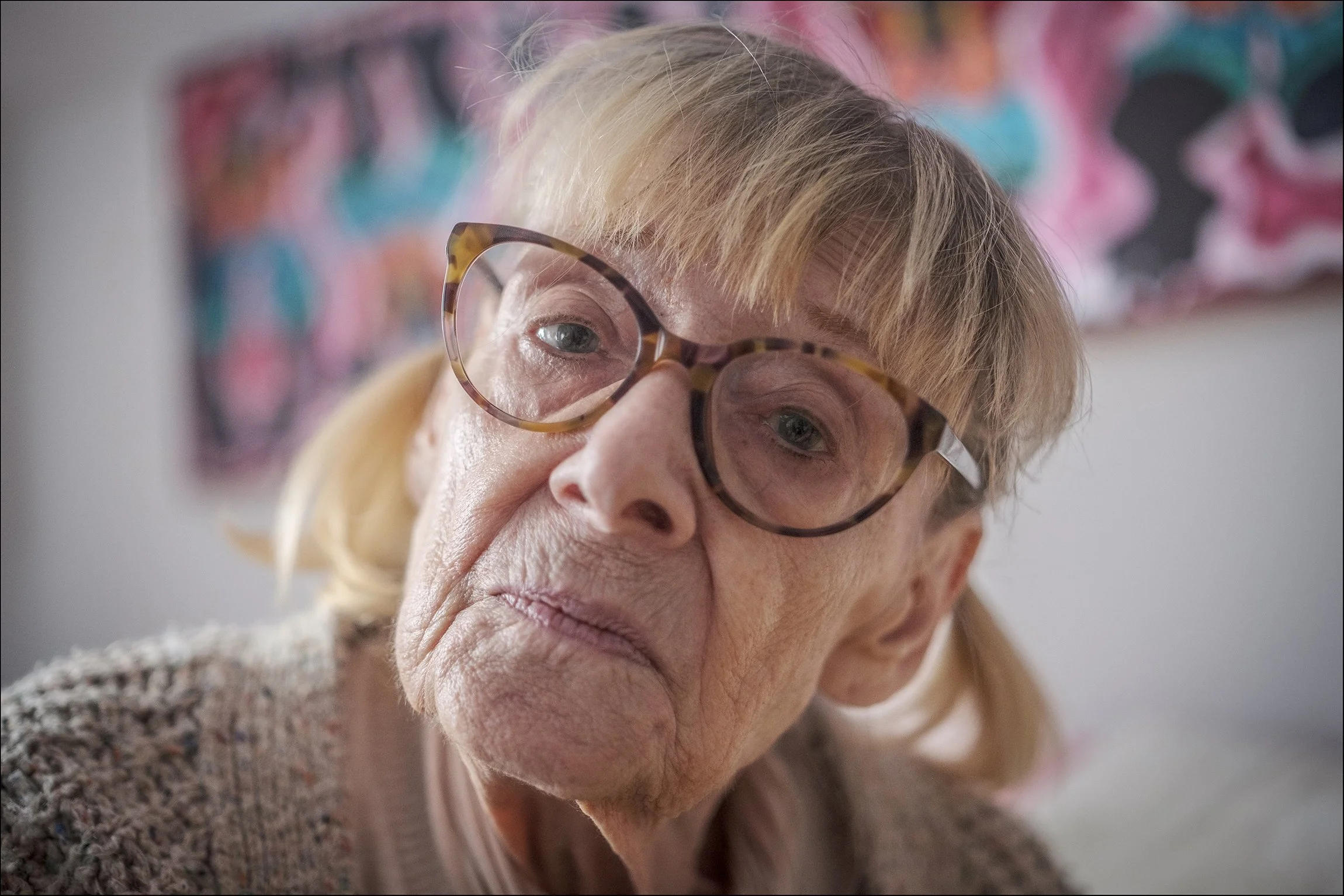 Nahaufnahme einer älteren Frau mit Brille, die einen nachdenklichen Ausdruck hat, im Hintergrund ein buntes Gemälde.
