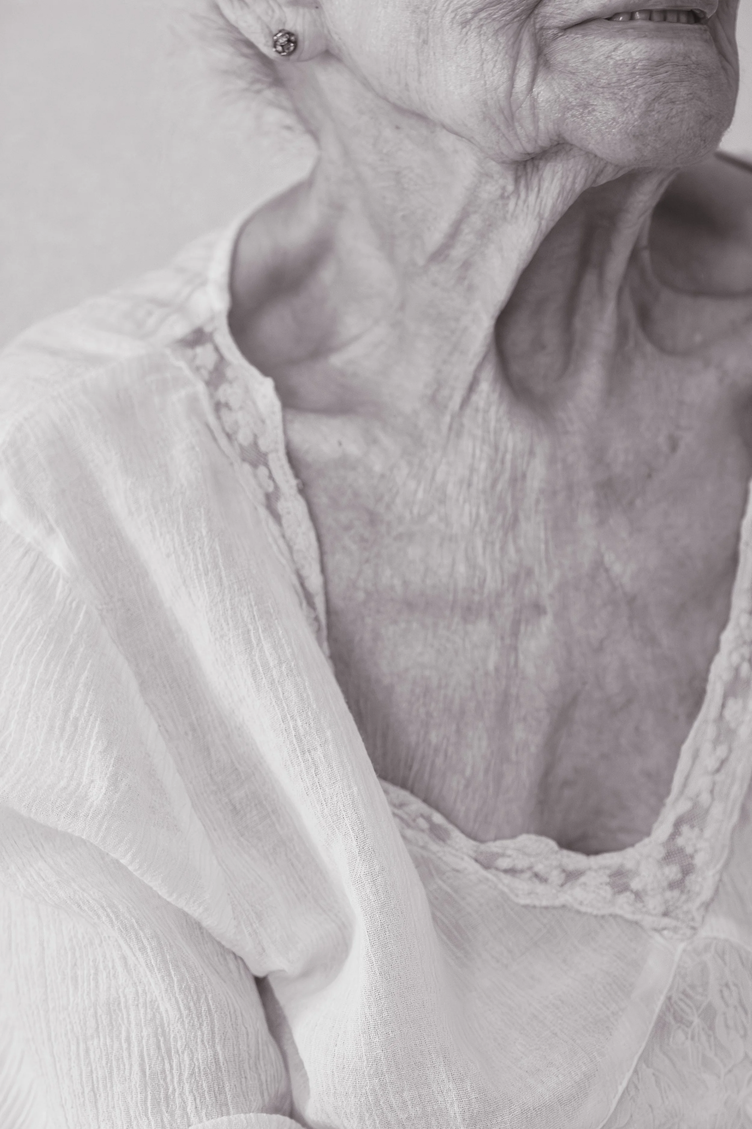 Eine Nahaufnahme des Halses und Teils des Gesichts einer alten Frau, bei der man die feinen Linien, Hauttextur und eine kleine Ohr-Perlen-Schmuckstück sieht. Sie trägt ein weißes Kleid mit Spitzenbesatz.