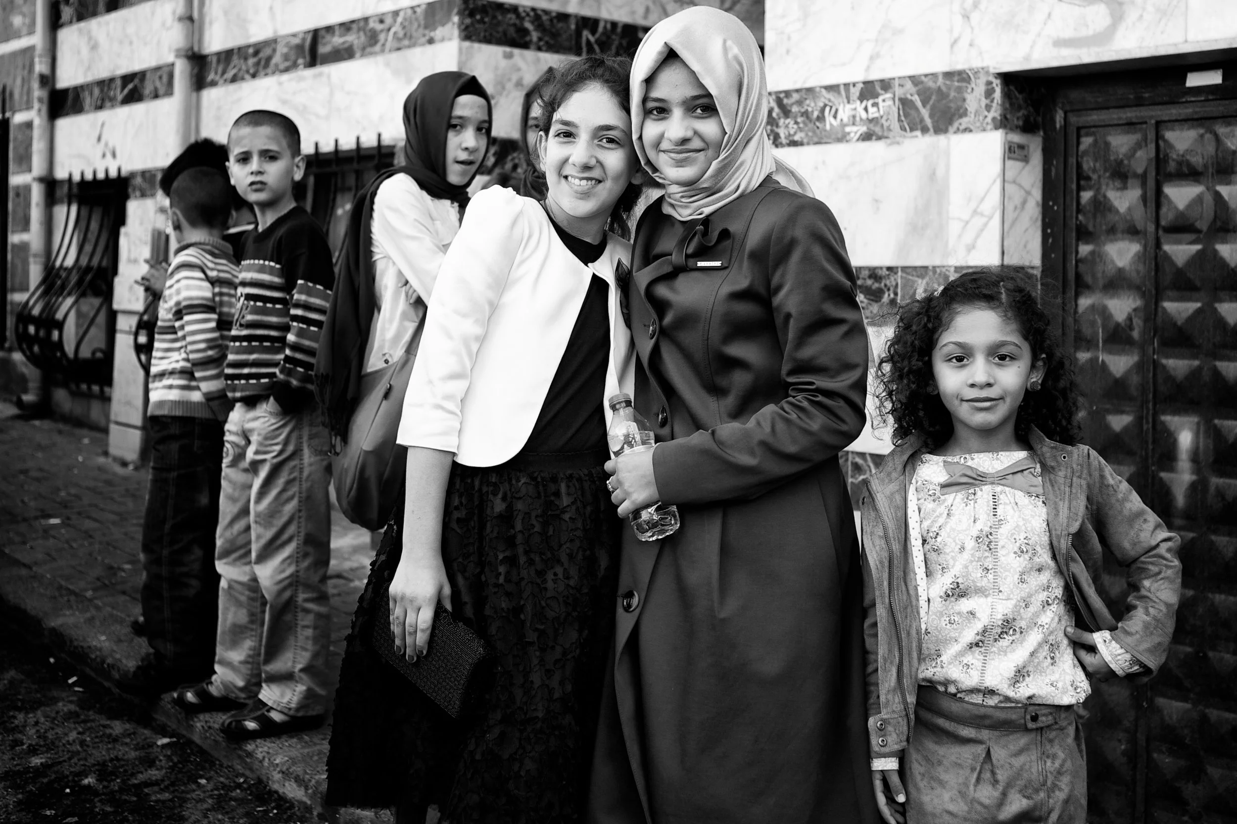 Vier Kinder stehen vor einer Wand aus Marmorfliesen, zwei Mädchen in Vordergrund und zwei Jungen im Hintergrund, eines der Mädchen trägt einen Hijab, alle Kinder schauen in die Kamera.
