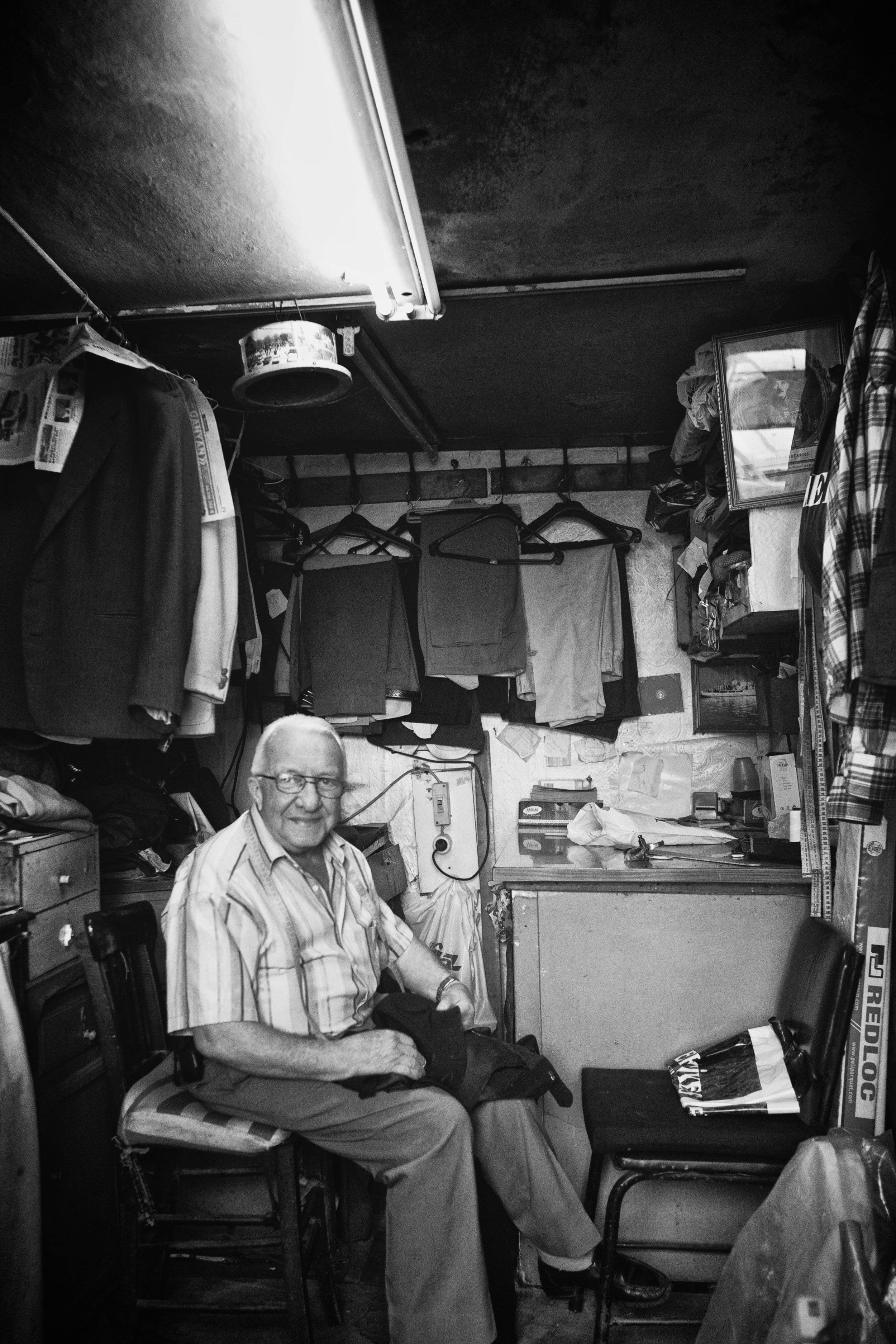 Ein älterer Mann sitzt in einem kleinen, chaotischen Raum voller Kleidung und allerlei Gegenstände, in schwarz-weiß.