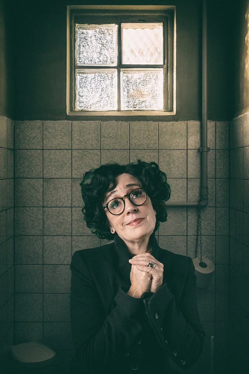 Eine Frau mit Brille in einem Raum mit Fliesen an der Wand, vor einer kleinen Fenster mit Eisblumen. Sie trägt schwarze Kleidung und schaut nach oben.