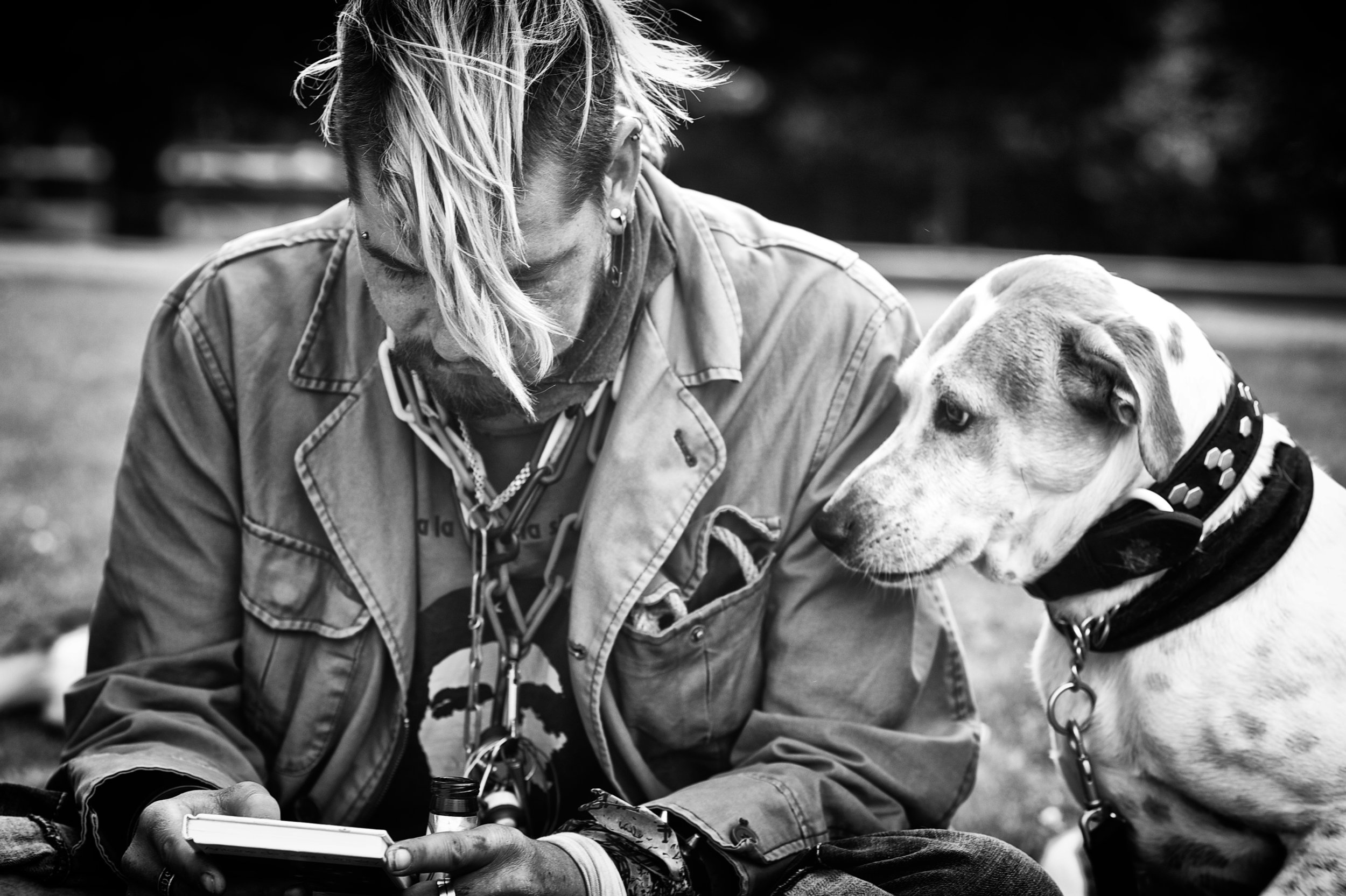 Ein Mann mit einem Dreadlock-Haarschnitt und mehreren Halsketten sitzt im Freien, hält ein Smartphone und schaut zu einem Dalmatiner-Hund. Das Bild ist schwarz-weiß.