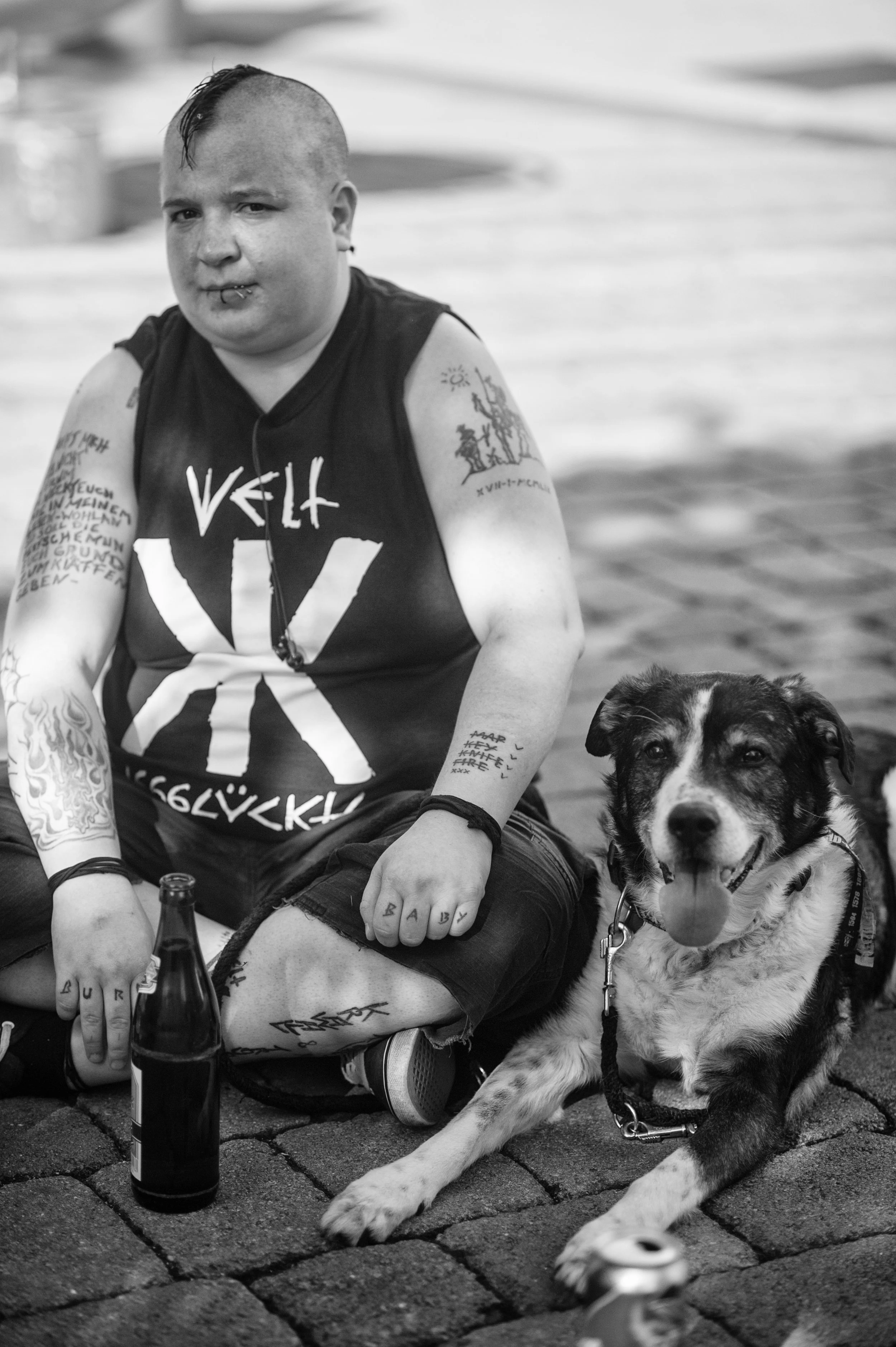Ein junge Frau mit Tattoos und Lippenpiercing sitzt auf Kopfsteinpflaster neben einem Hund
