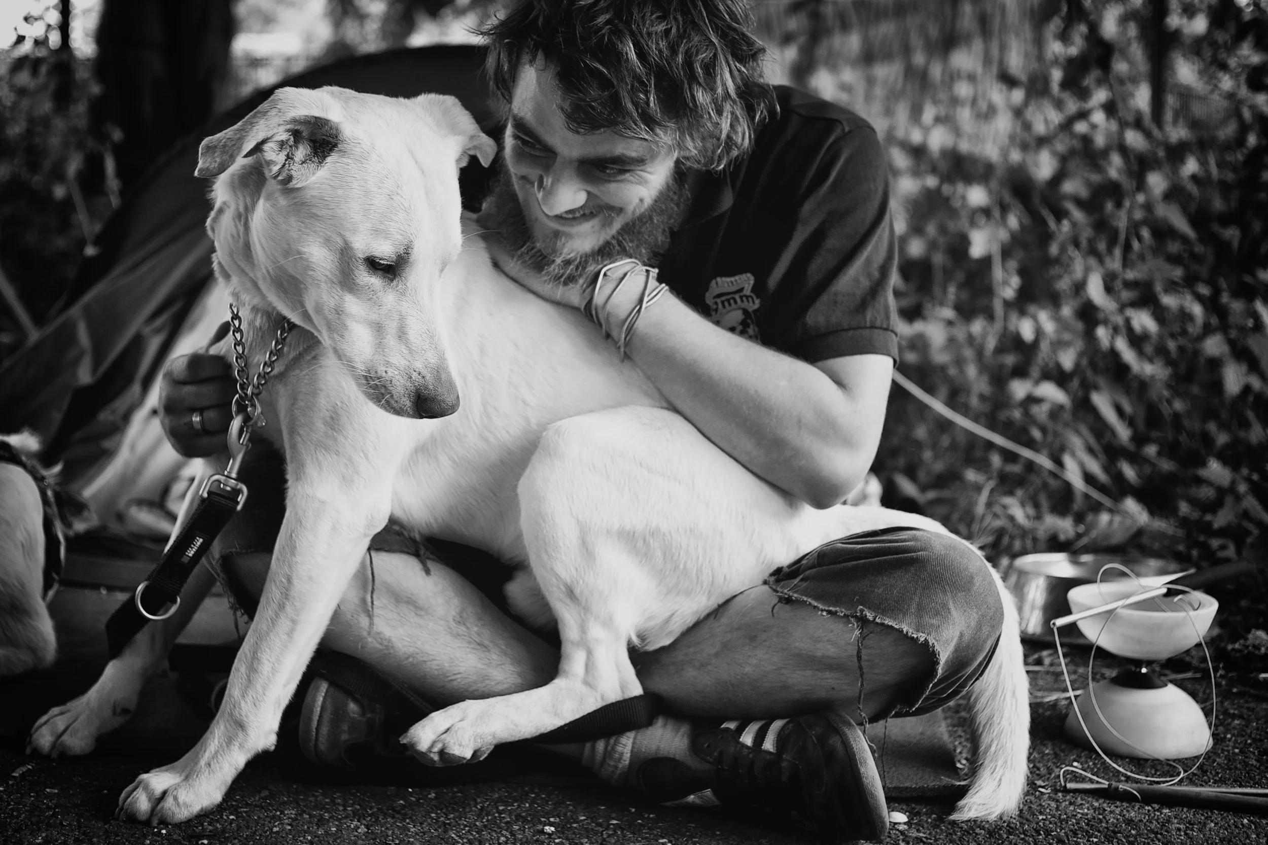 Ein Mann sitzt im Freien auf dem Boden mit einem weißen Hund auf seinem Schoß, beide scheinen sich zu freuen. Es ist schwarz-weiß Foto.