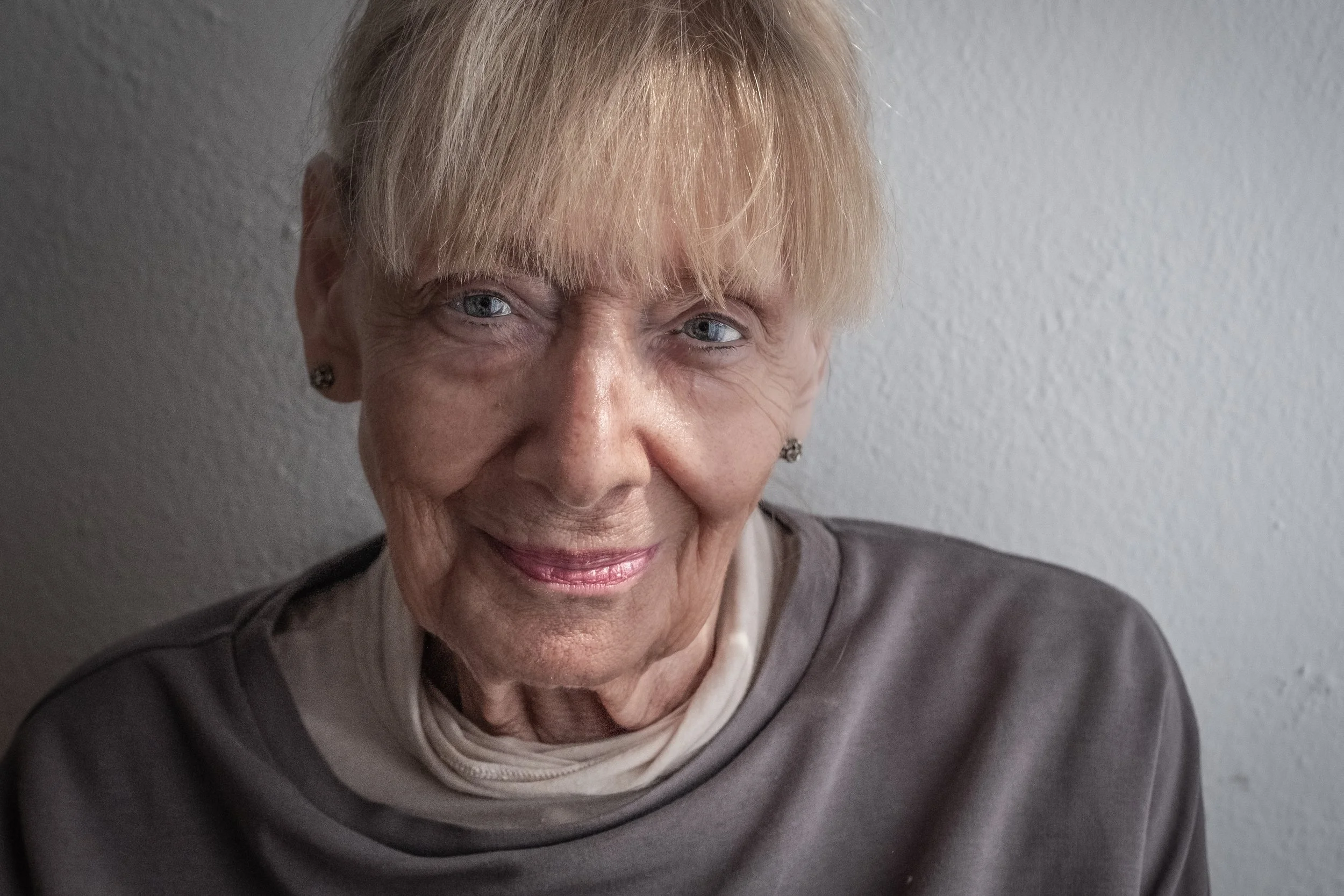 Nahaufnahme einer älteren Frau mit blondem Haar, blauen Augen und einem freundlichen Lächeln vor einer hellgrauen Wand.