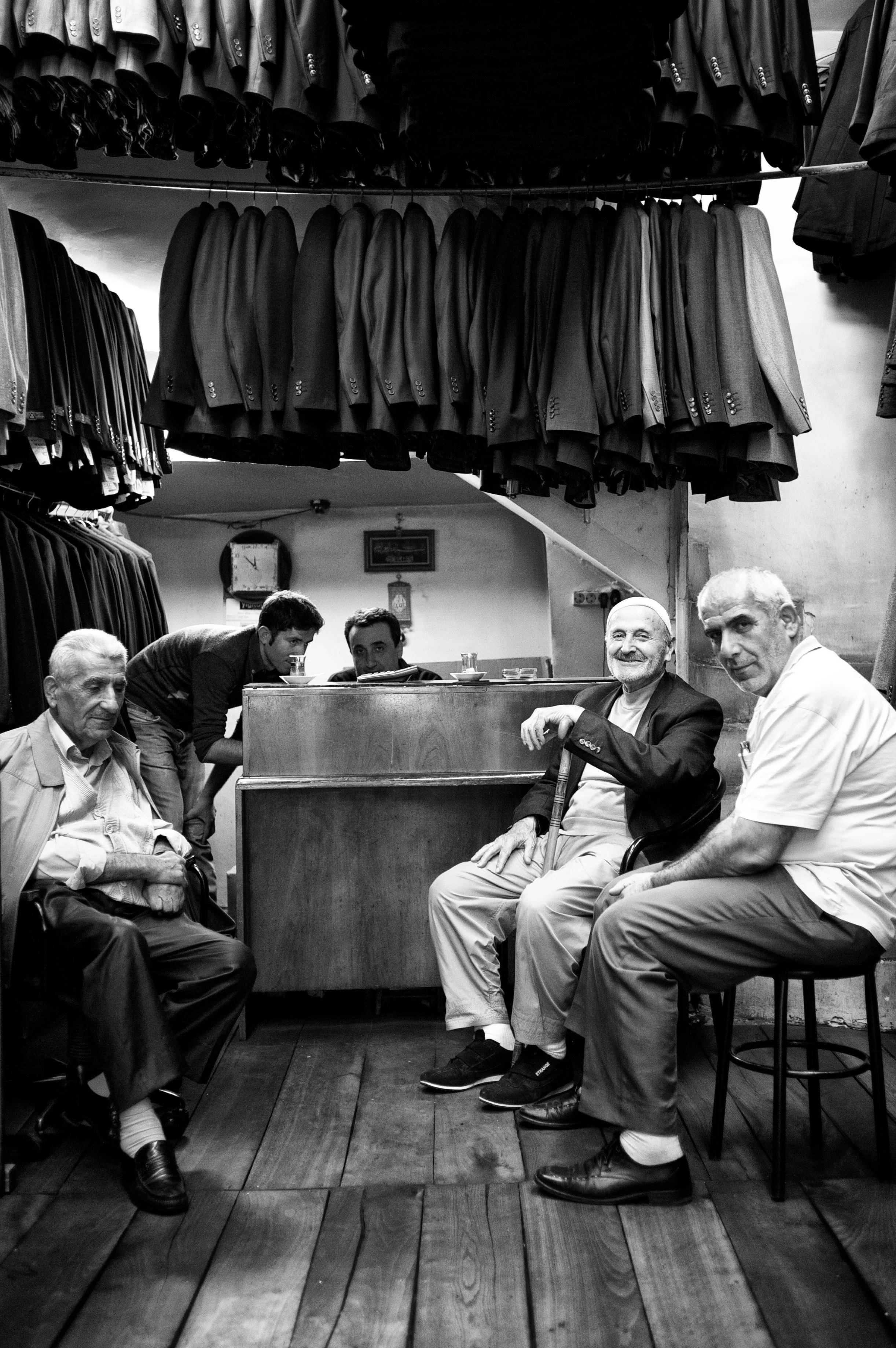 Vier Männer sitzen in einem kleinen Laden, im Hintergrund hängen Anzüge. Zwei junge Männer stehen hinter einem Holztresen, der in der Mitte im Bild sichtbar ist.