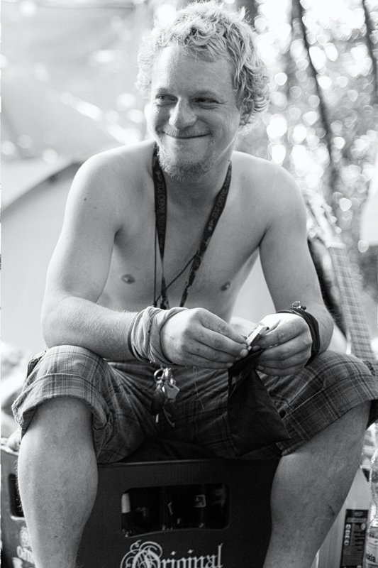 Locker lachender Mann sitzt barfuß auf einem Bierkasten im Freien, trägt eine Halskette, eine Armband und ein Fahrrad im Hintergrund.