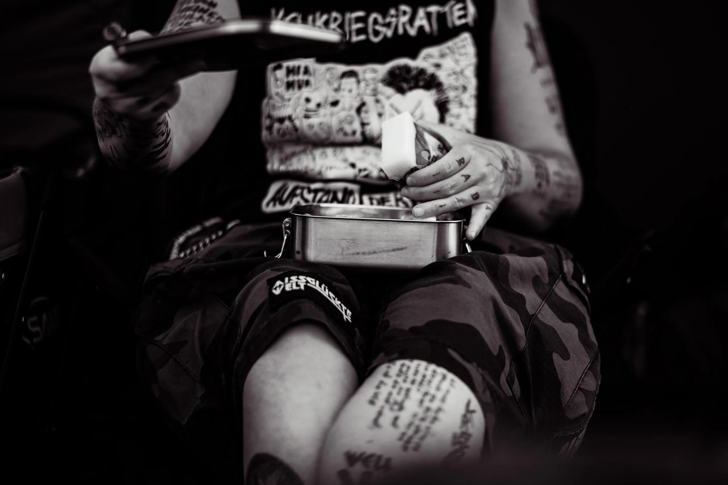 Nahaufnahme eines Tattoospezifischen Oberschenkels, auf dem ein Riegel Schokolade auf einem Metallbehälter liegt, während eine Person mit tätowierten Armen einen Bissen Schokolade hält, in Schwarz-Weiß-Fotografie.