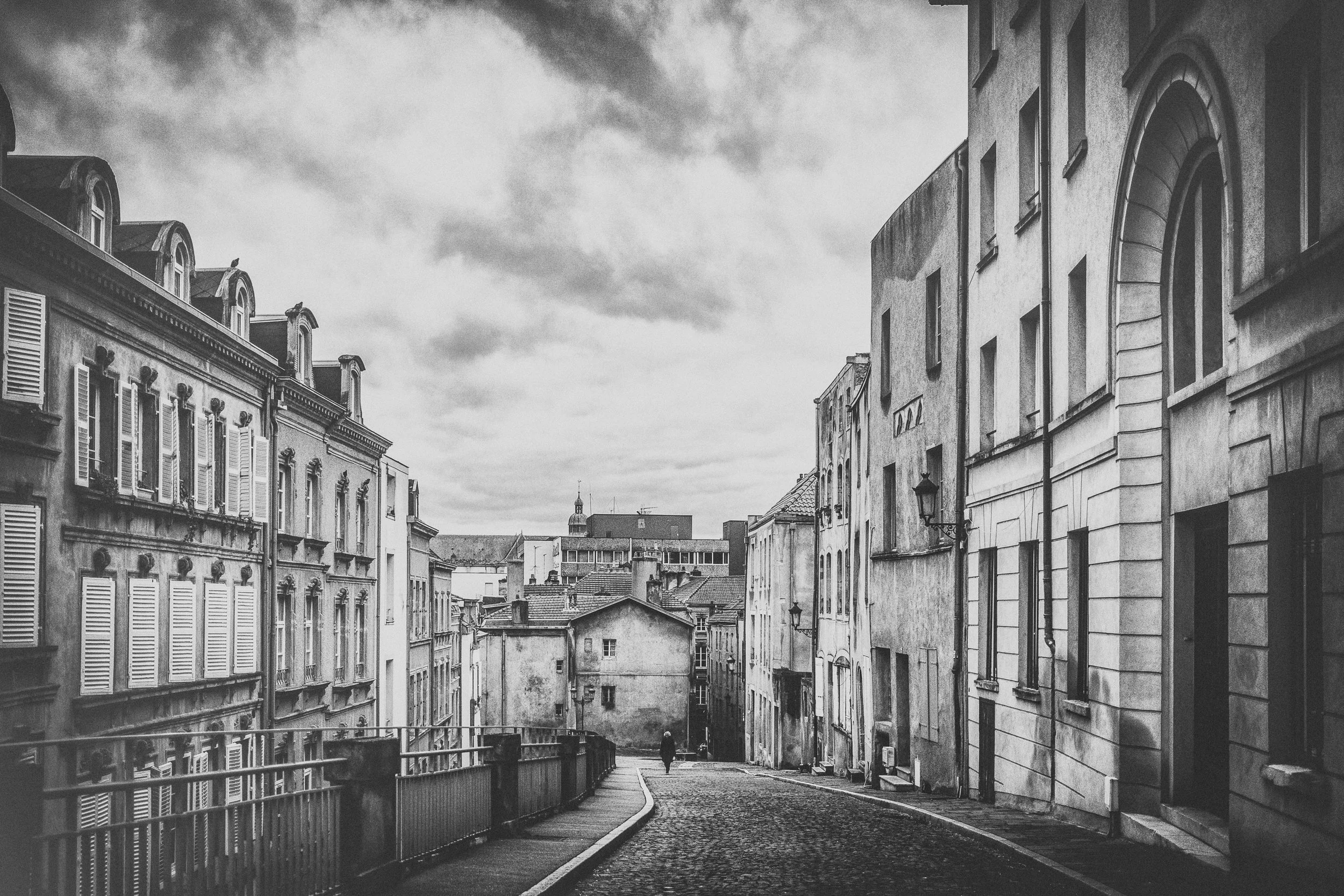 Eine leere, schmale, gewundene Straße in einer Stadt mit alten Gebäuden und Wolken am Himmel, in Schwarz-Weiß.