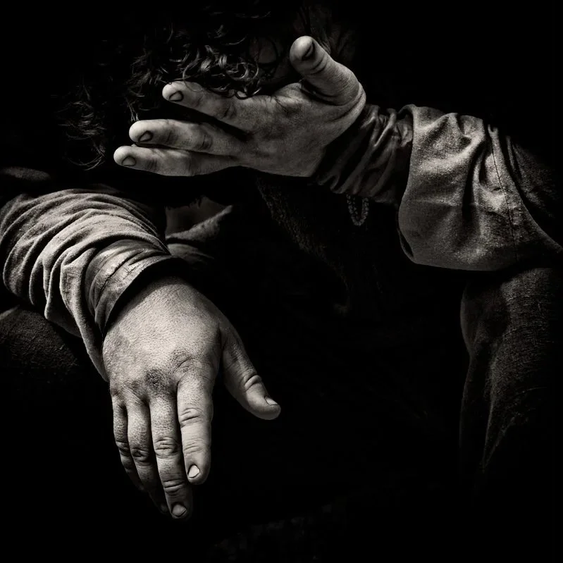 Schwarze und weiße Nahaufnahme von einer Person, die ihre Hände auf das Gesicht legt und den Kopf neigt, mit sichtbaren, schmutzigen Händen und getragenen Kleidung.