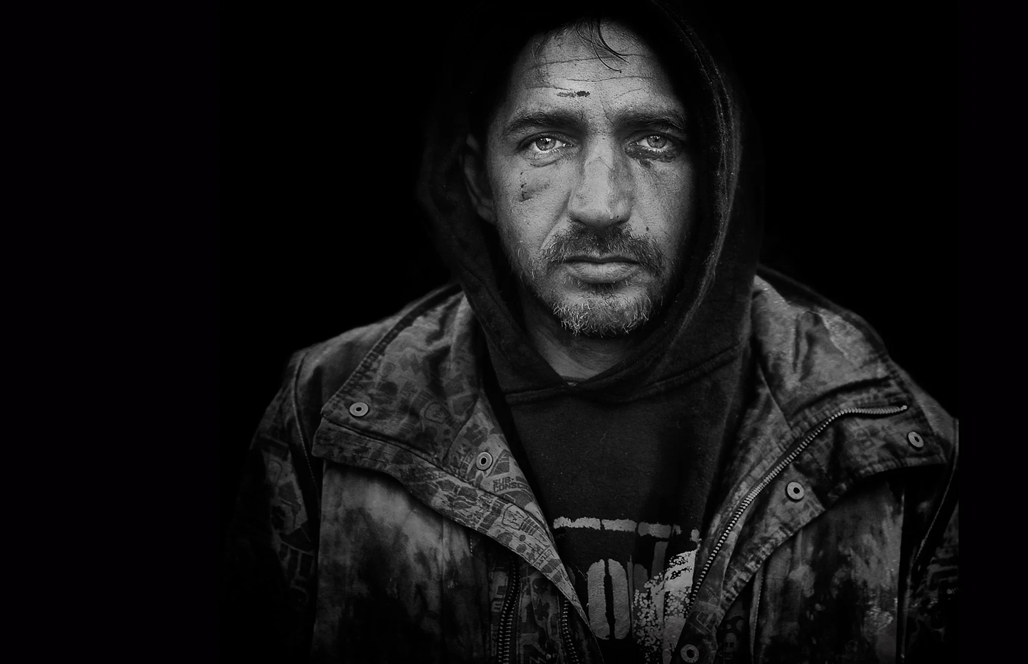 schwarz-weiß Porträt eines Mannes mit zerzaustem Bart und schwarzen Haaren, der eine dunkle Kapuzenjacke trägt, vor dunklem Hintergrund.