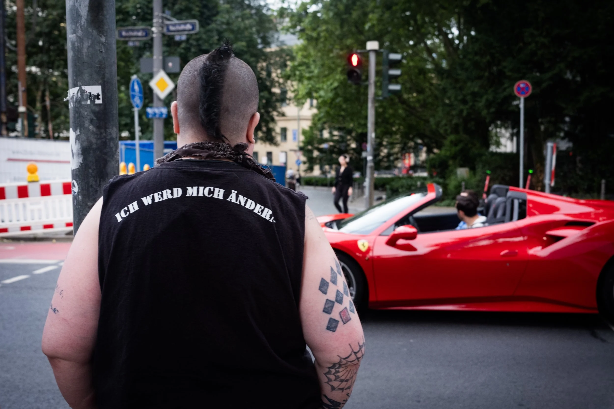 Person mit tätowierter Arm und schwarzem T-Shirt mit weißen Aufdruck, Blick auf rote Ferrari-Sportwagen vor einer Kreuzung in der Stadt.