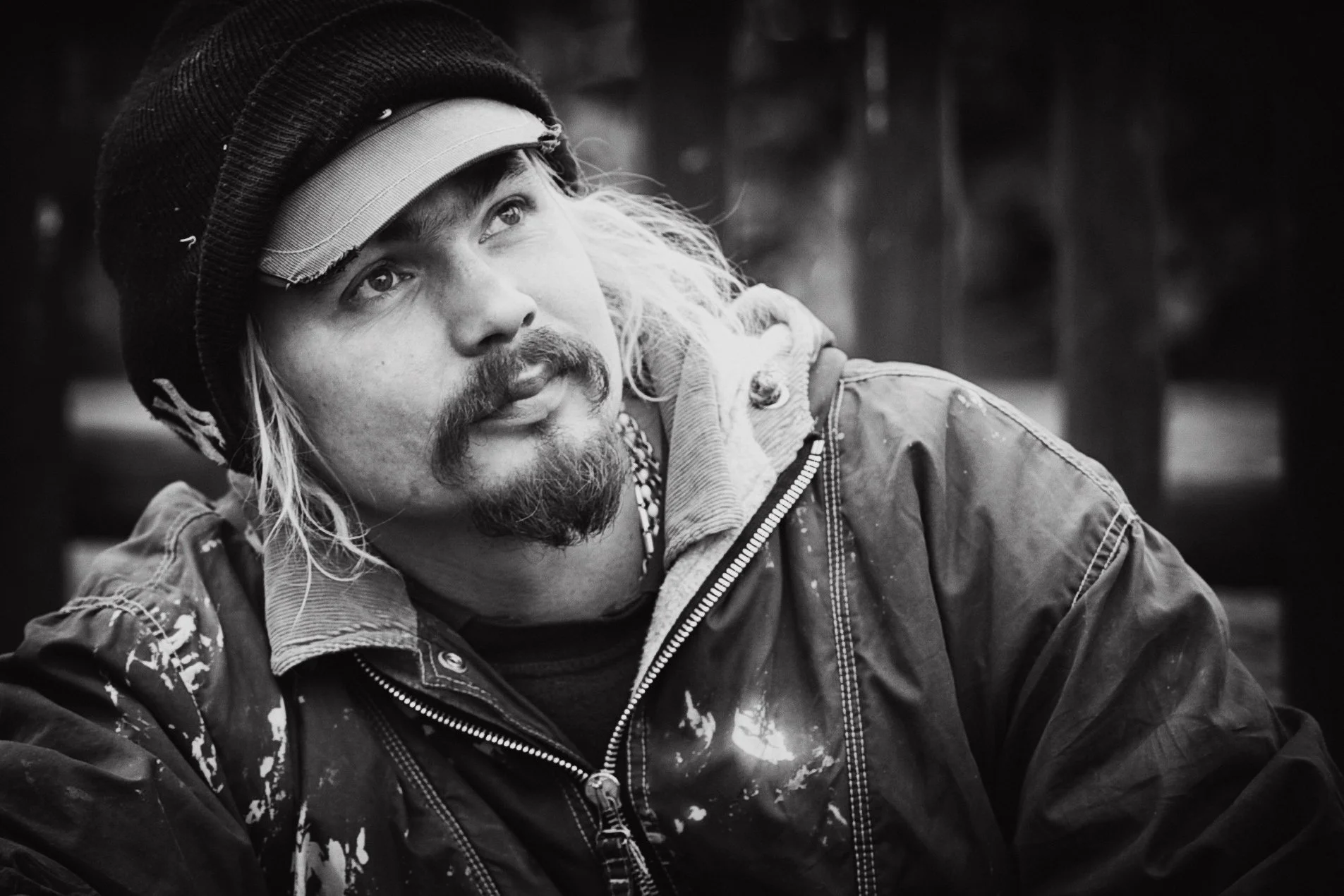 Schwarz-Weiß-Fotografie eines Mannes mit Helm, Kinnbart und langen Haaren, der nach oben schaut, trägt eine wetterfeste Jacke mit deutlichen Abnutzungen.