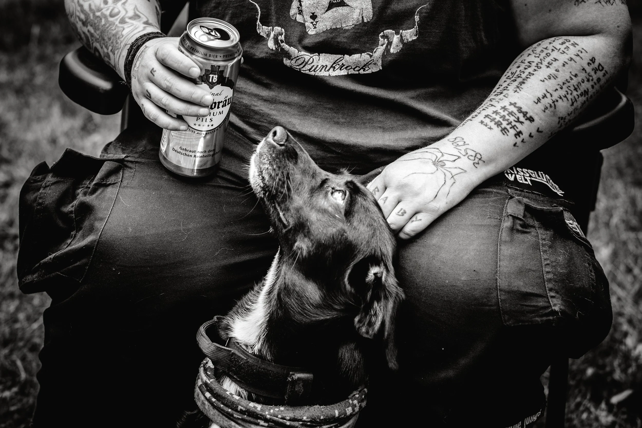 Eine Person sitzt mit einer Bierdose auf seinem Schoß, während ein Hund an seiner Seite nach oben schaut. Die Person hat tätowierte Arme und trägt eine schwarze Hose. Das Bild ist schwarz-weiß, und der Hintergrund ist unscharf.