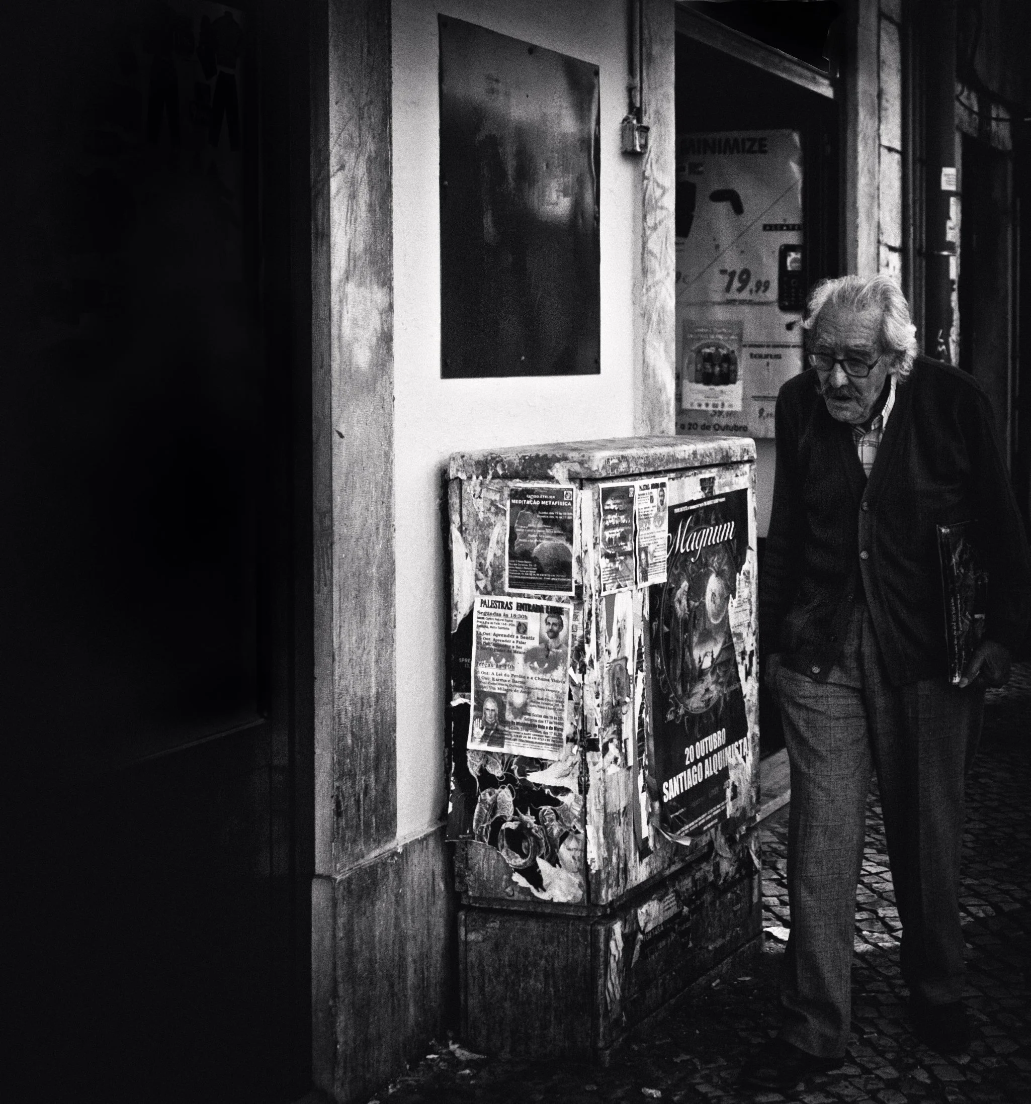 Ein älterer Mann mit Brille und dunkler Jacke liest eine Info-Tafel auf einem Straßenrand in einer urbanen Gegend, schwarz-weiß Foto.