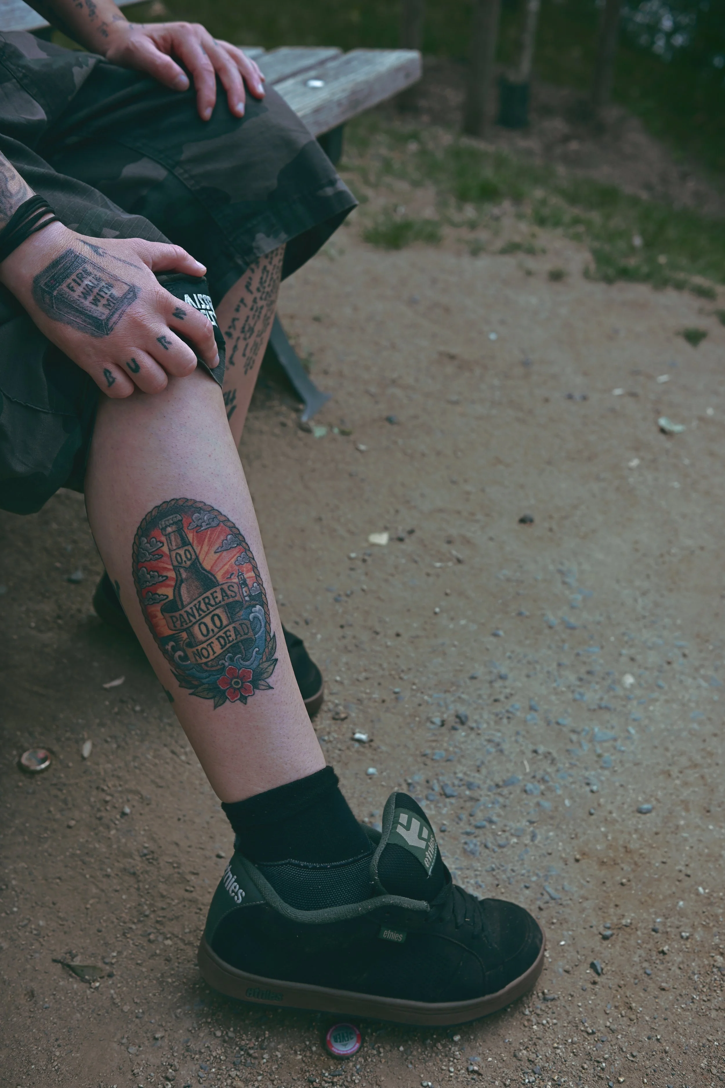 Person sitzt auf einer Bank, zeigt eine Tattoo auf dem Unterschenkel mit der Aufschrift "PANKREAS 0.0 NOT DEAD" und einem Flaschenmotiv, trägt schwarze Schuhe und eine schwarze Socke.
