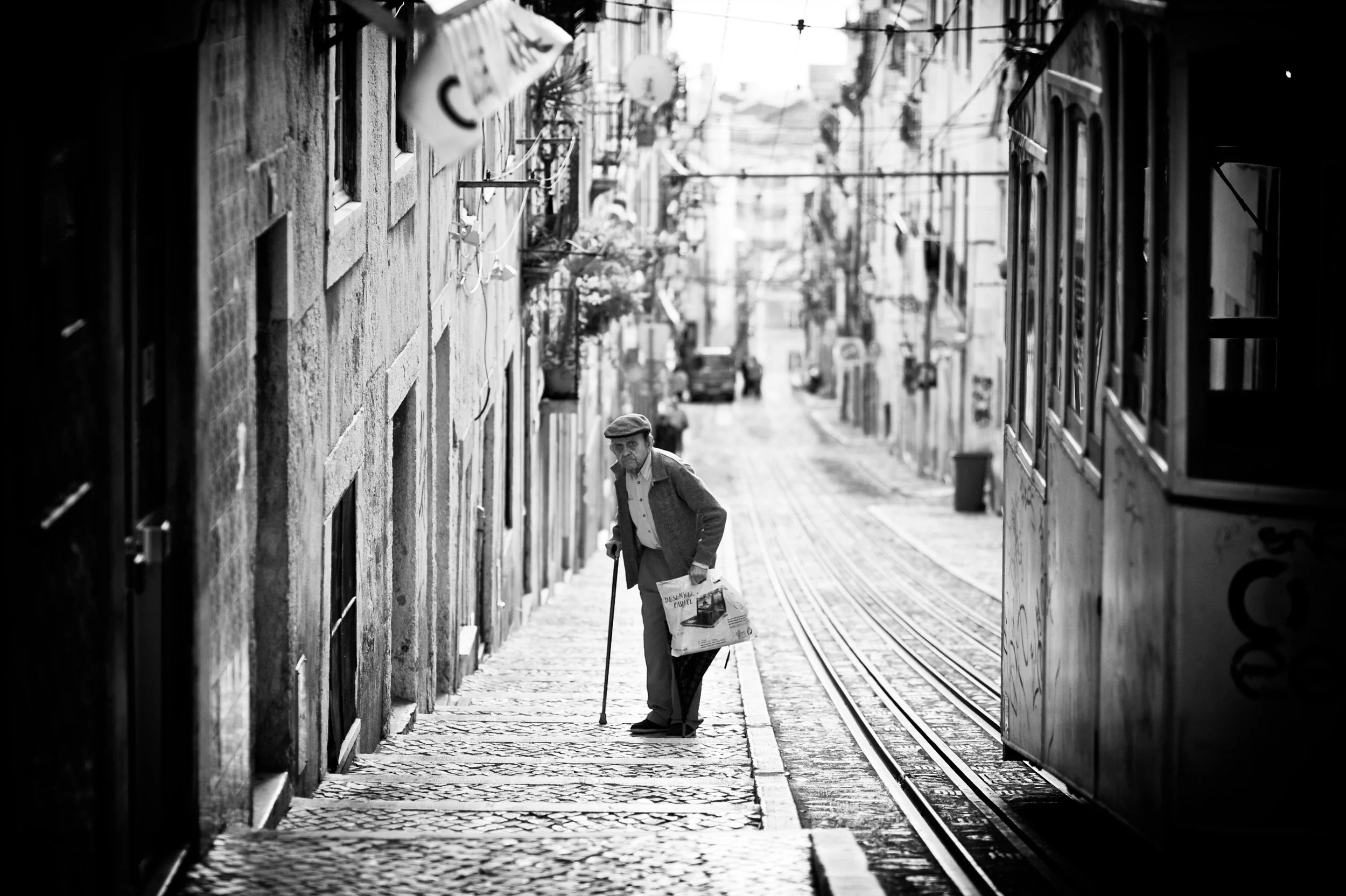Alter Mann mit Stock und Einkaufstüte auf einem gepflasterten Bürgersteig neben Straßenbahn in einer schmalen, hügeligen Stadtstraße, schwarz-weiß