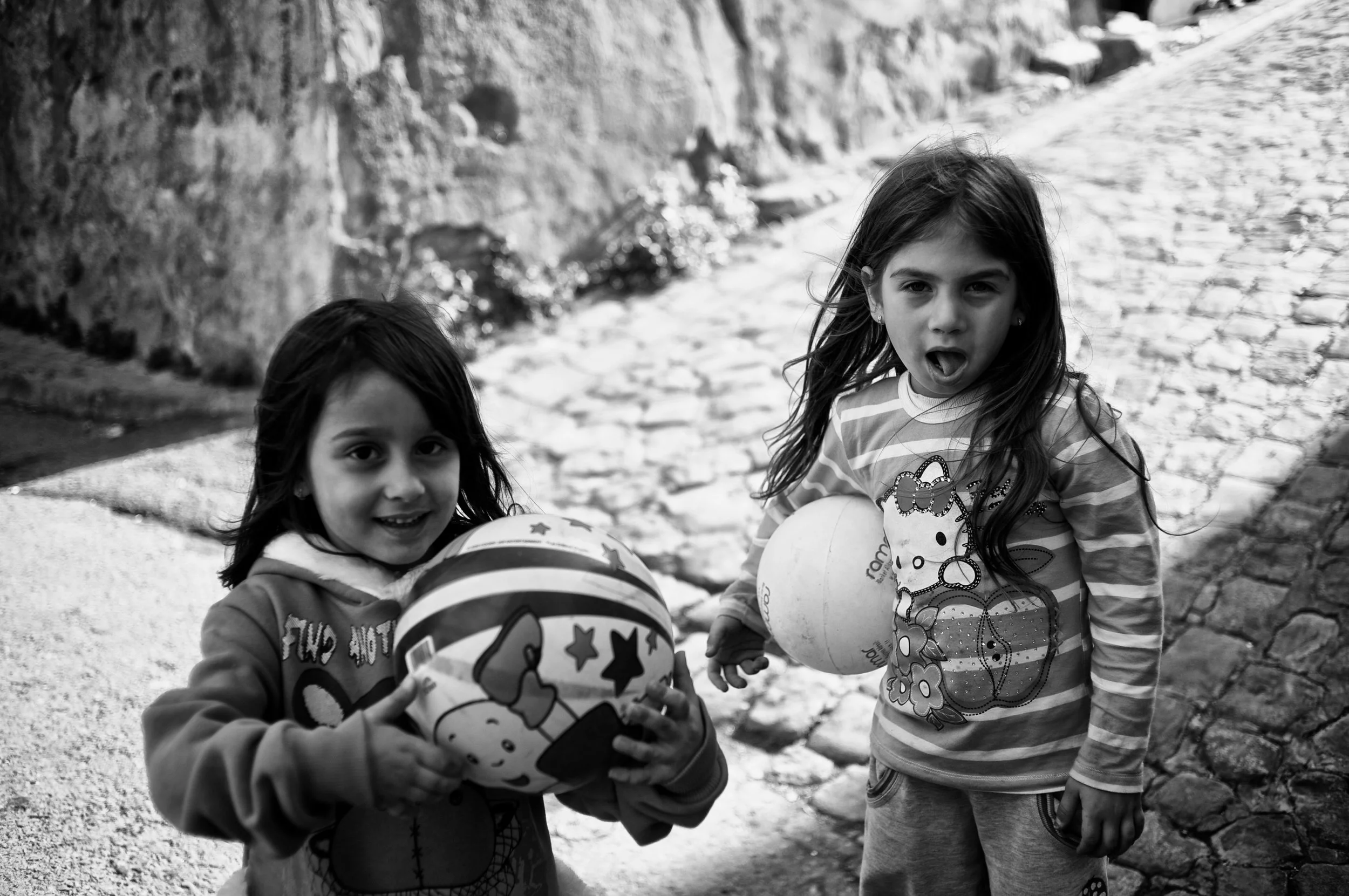 Zwei junge Mädchen stehen auf einer gepflasterten Straße, eines hält einen Ball mit Hello Kitty Design, das andere einen weißen Ball. Beide Mädchen schauen in die Kamera.