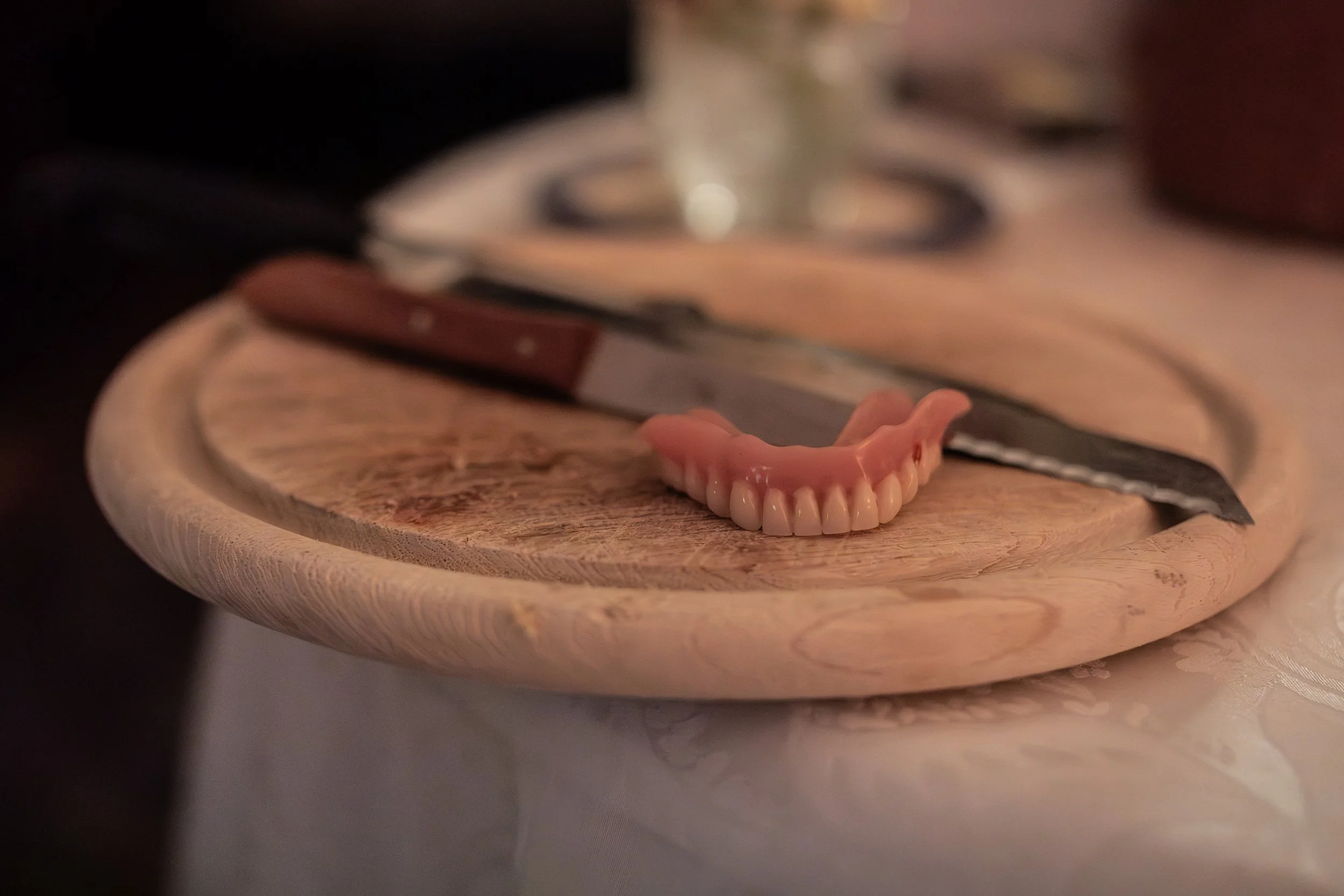 Holzbrett mit einem Küchenmesser, Simulationszahnset und einem Glas. Die Szene wirkt wie eine Küchen-oder Essenszubereitung.