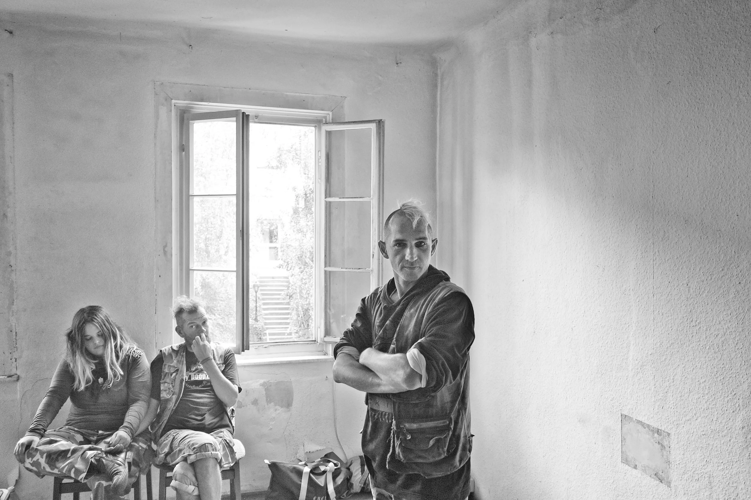 Drei junge Menschen sitzen in einem Raum mit offener Fenster. Einer steht im Vordergrund und schaut zur Seite, während die anderen zwei im Hintergrund sitzen. Es ist ein Schwarz-Weiß-Foto.