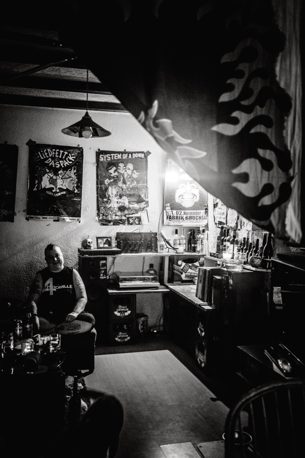 Ein dunkler, schwarz-weißes Foto eines gemütlichen Raums mit einer Person, die auf einem Stuhl sitzt und mehrere Getränke auf einem Tisch vor sich hat. Die Wand im Hintergrund ist mit Postern und Fotos dekoriert, darunter ein System of a Down Poster.