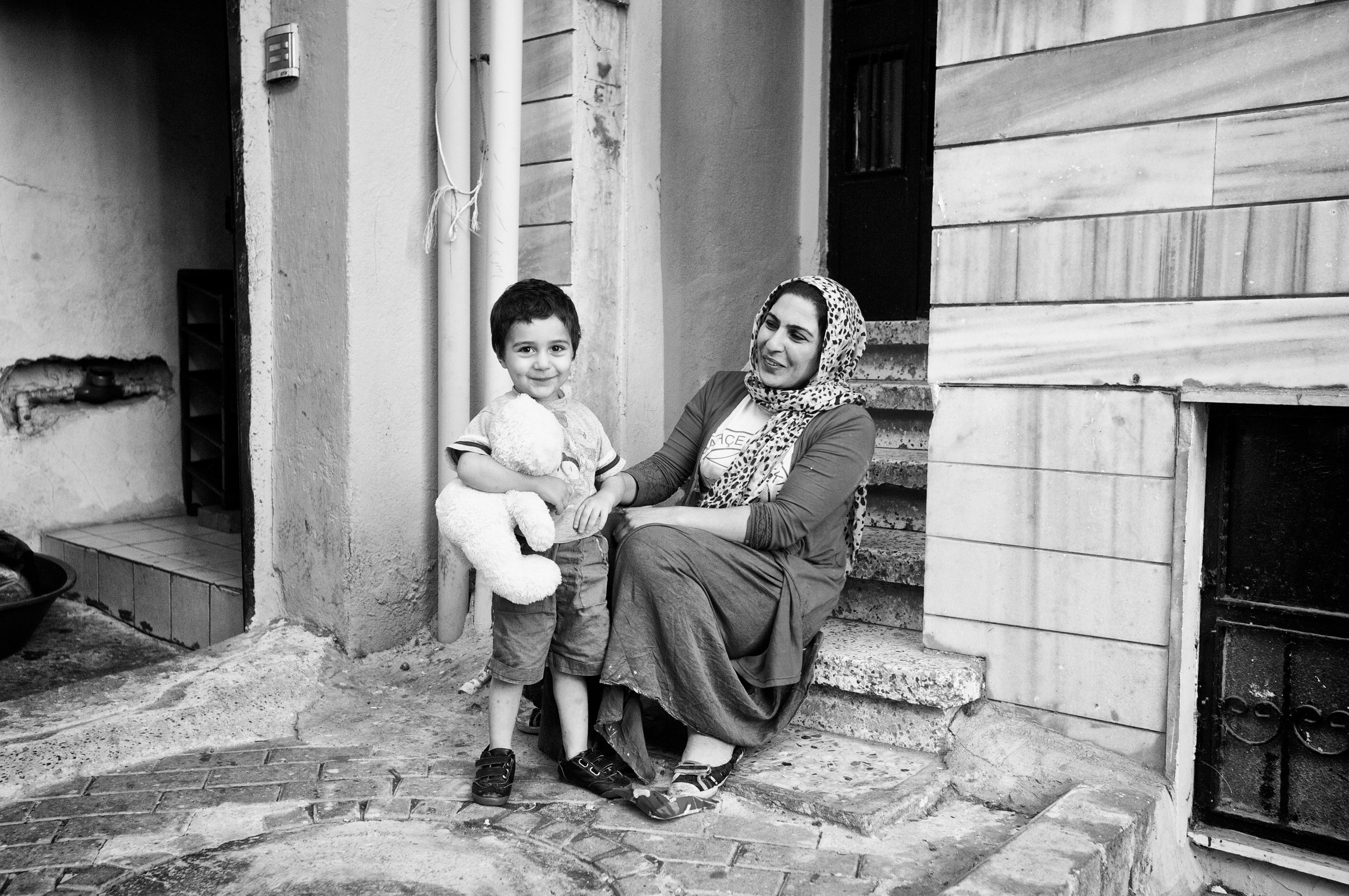 Ein lachendes kleines Mädchen mit einem Teddybären steht vor einer Frau, die auf Steinstufen sitzt, vor einem Haus mit Holzverkleidung. Es ist schwarz-weiß.