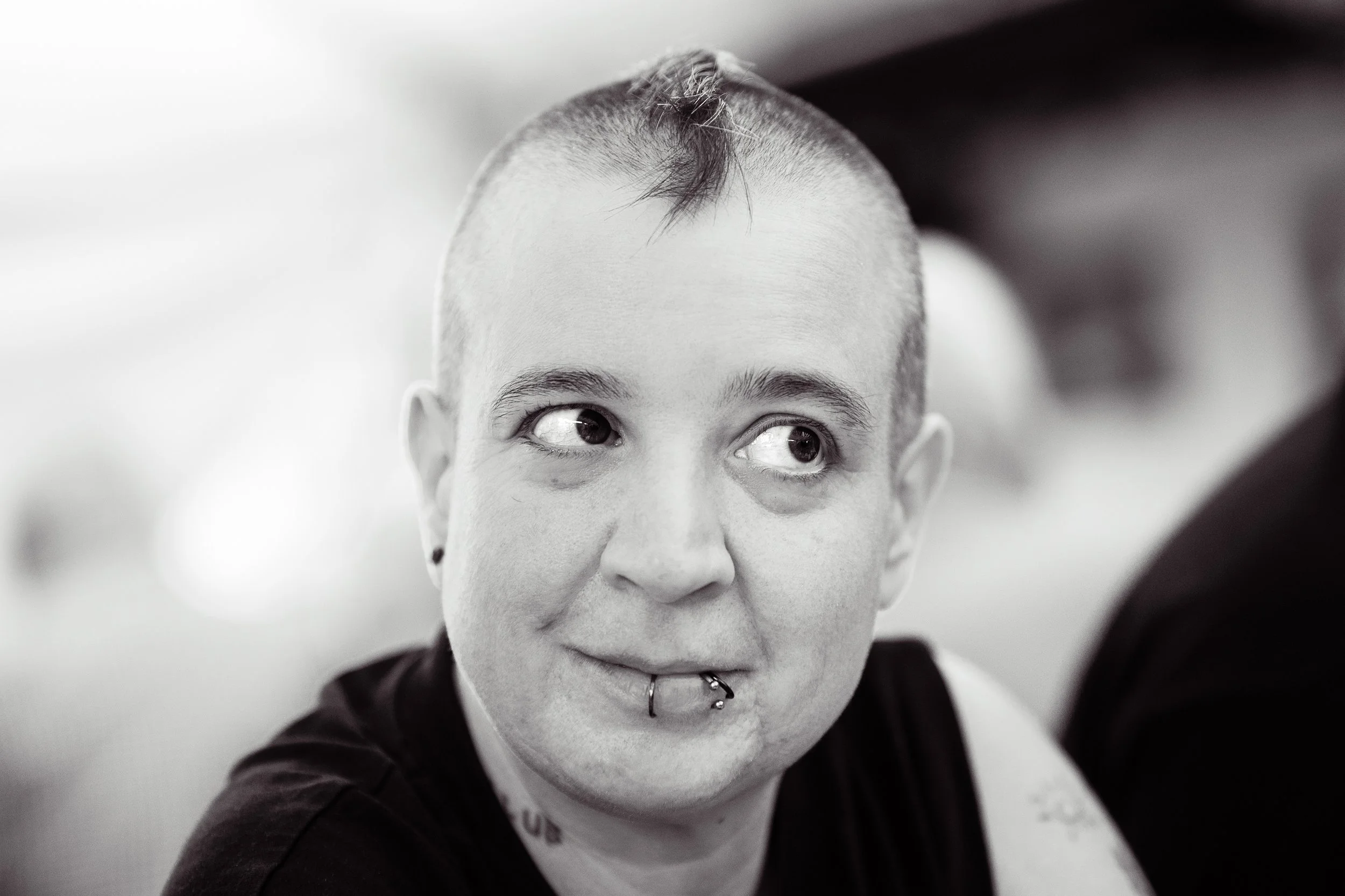 Schwarz-weiß-Fotografie eines jungen Menschen mit kurzen Haaren, Piercing im Mund, schwarzen Ohrringen und Tattoo am Arm, der leicht zur Seite schaut.