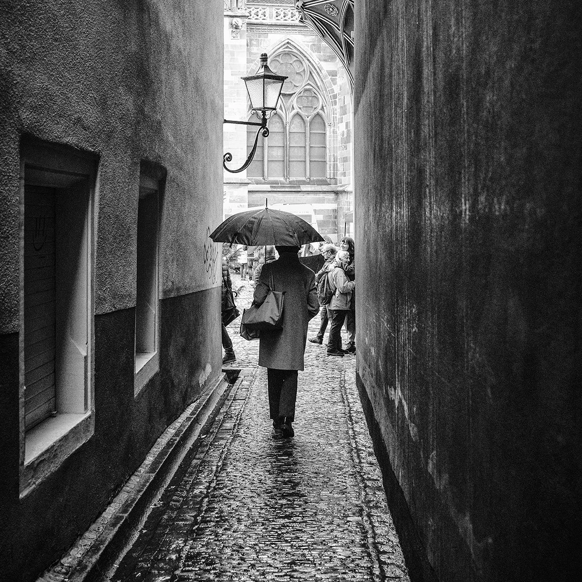 Person von hinten, die mit einen Regenschirm durch eine enge Gasse geht. Am Ende der Gasse stehen 2 Personen, die sich umarmen