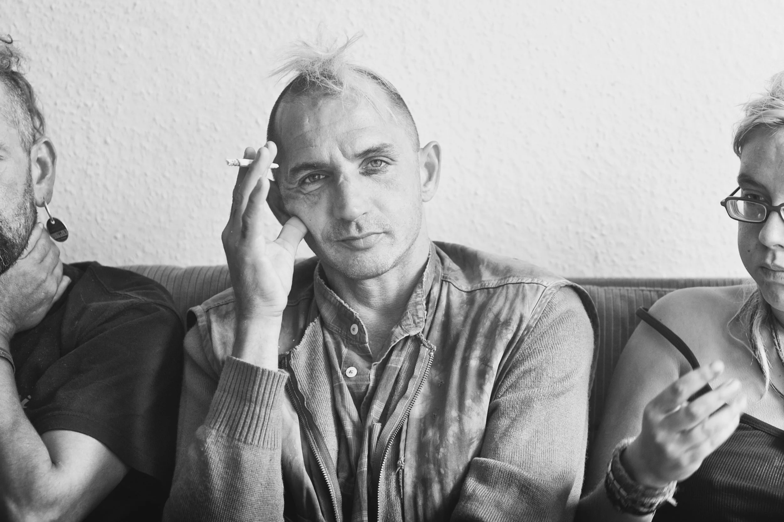 Schwarz-weiß-Foto von drei Personen sitzend auf einer Couch, der mittlere Mann raucht, trägt einen gesteppten Mantel, hat kurz geschnittenes Haar und blickt direkt in die Kamera.