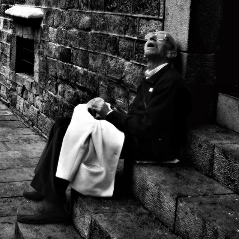 Ein alter Mann sitzt erschöpft auf einer Treppenstufe gegen eine Steinmauer, die Augen geschlossen, scheint zu schlafen oder sich auszuruhen.