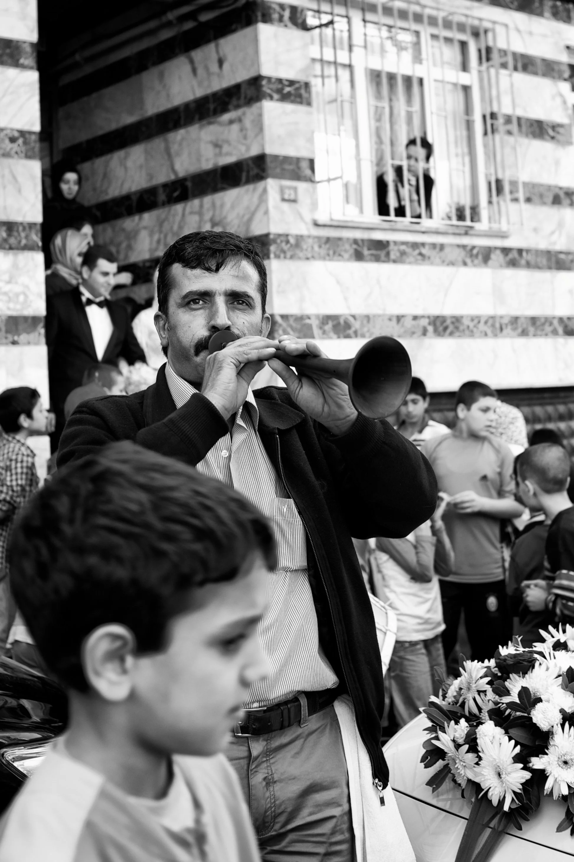 Mann spielt auf einer Flöte in einer Menschenmenge bei einer Veranstaltung, im Hintergrund eine Person, die aus einem Fenster schaut, und Kinder, die um Blumen stehen.
