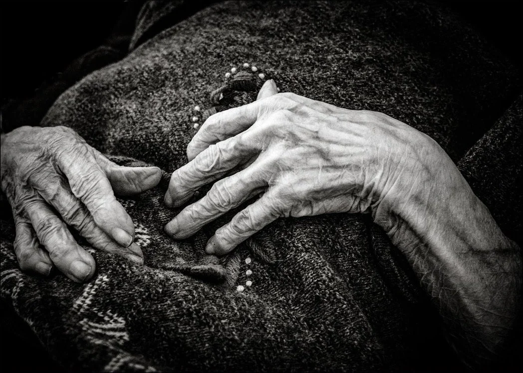 Gealterte, knochige Hände in schwarz-weiss. Sie liegen auf einer teilweise bestickten Decke, vermutlich auf der Brust der Person