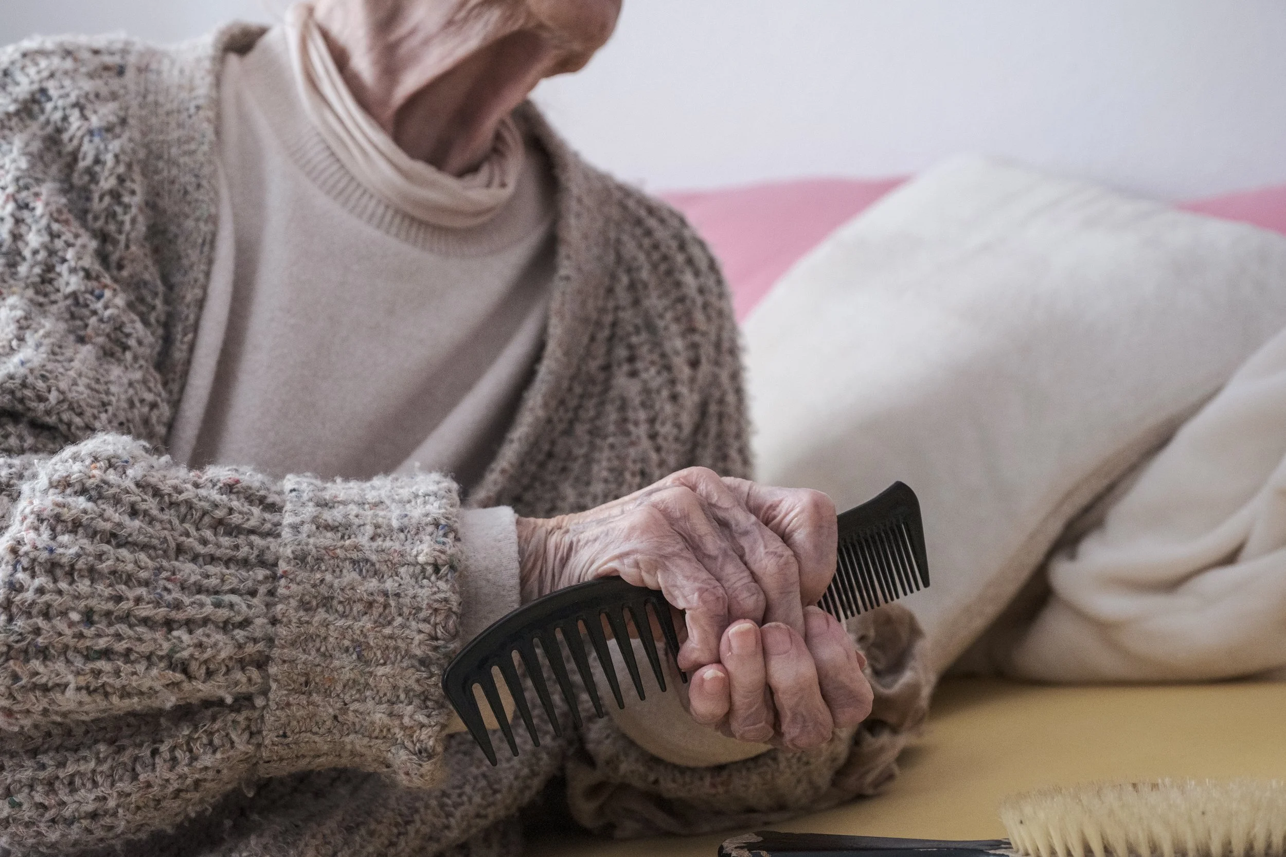 Eine ältere Person kämmt ihr Haar mit einem schwarzen Kamm auf einem Bett.