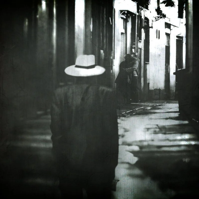 Schwarz-weiß-Foto einer straßen Szene mit einer Person, die gegen die Wand lehnt, und einer weiteren Person, die auf einem Gehweg läuft. Im Vordergrund ist ein Hut sichtbar, Reflexion eines Kopfes im Wasser auf dem Boden.