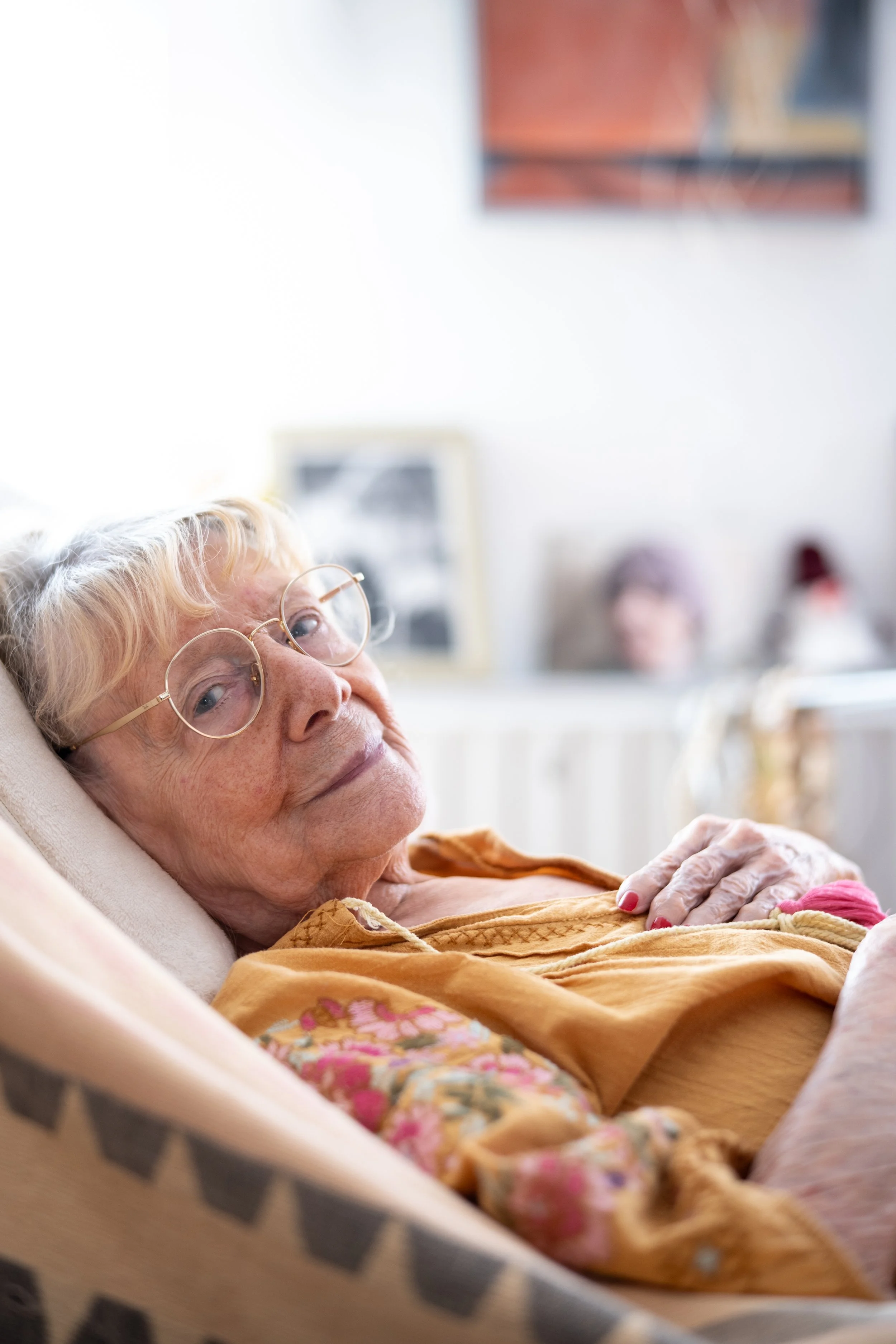 Ältere Frau mit Brille liegend auf einem Sofa, lächelnd, im Hintergrund Bilder und Dekoration in einem gemütlichen Raum.