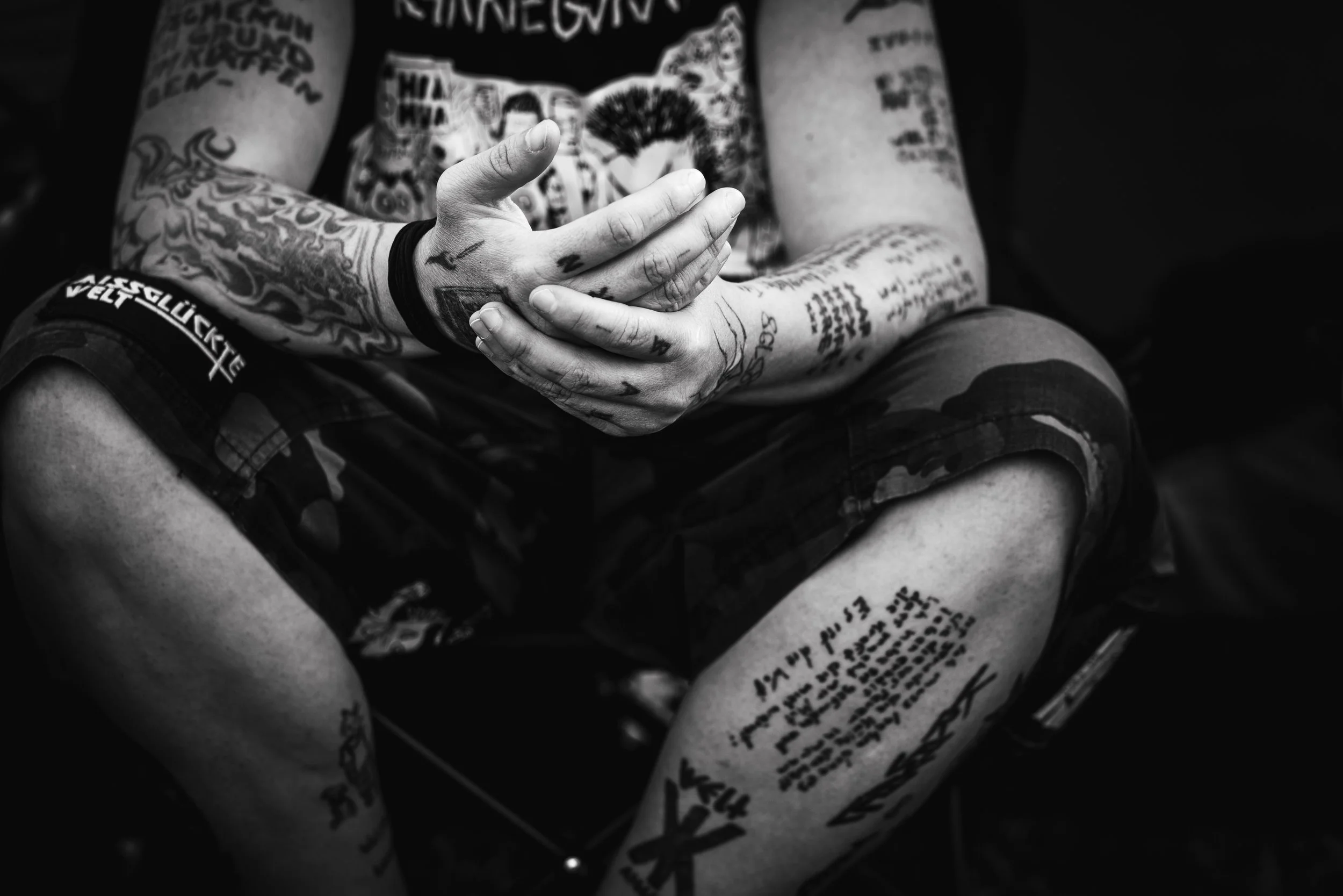 Ein Mann mit vielen Tattoos auf Armen, Händen und Beinen, der im Sitzen die Hände zusammenfasst. Er trägt eine schwarze Armband und eine kurze Hose mit Camouflagemuster. Das Bild ist schwarz-weiß.