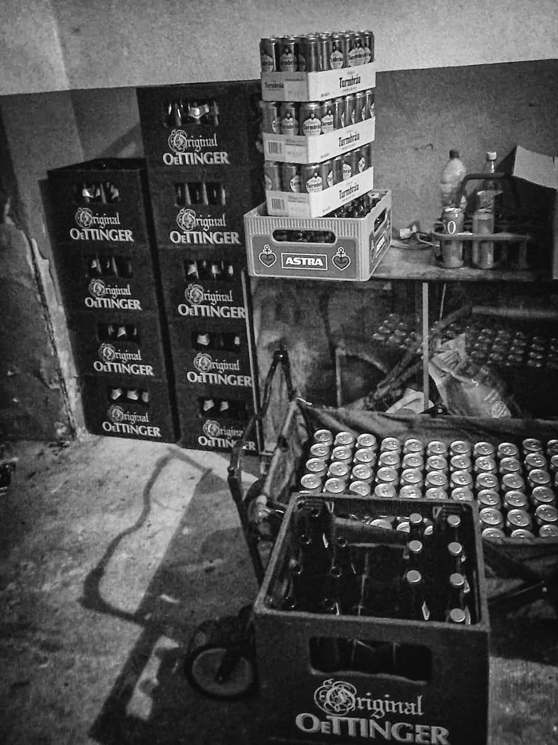 Büro- oder Lagerraum mit Getränkekisten, hauptsächlich Oettinger Bier, und einem Tisch mit weiteren Getränken, auf einem Stuhl liegt ein Kabel.