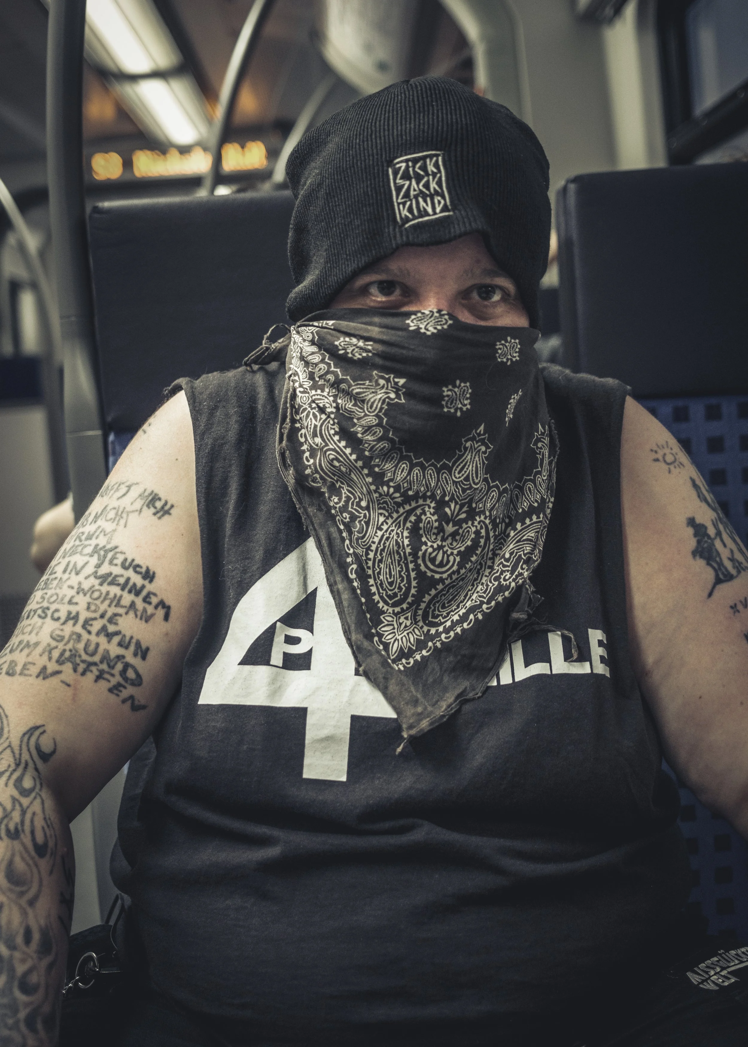 Ein Mann sitzt in einem Zug, trägt eine schwarze Mütze, ein Tuch vor dem Gesicht und ein schwarzes Tanktop mit einem weißen Symbol. Er hat Tätowierungen am Arm.