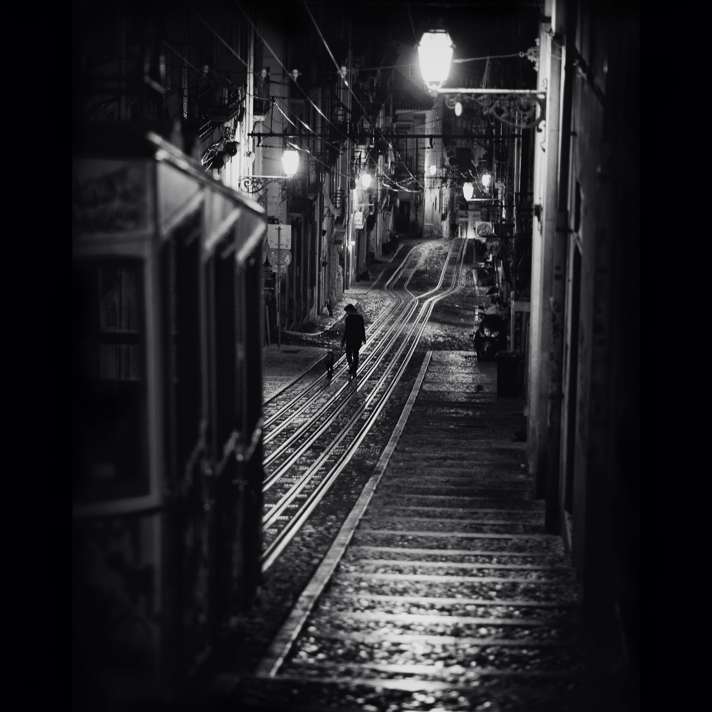 Eine nächtliche, beleuchtete Straße mit Schienen und einer einzelnen Person, die auf den Schienen geht, in Schwarz-Weiß.