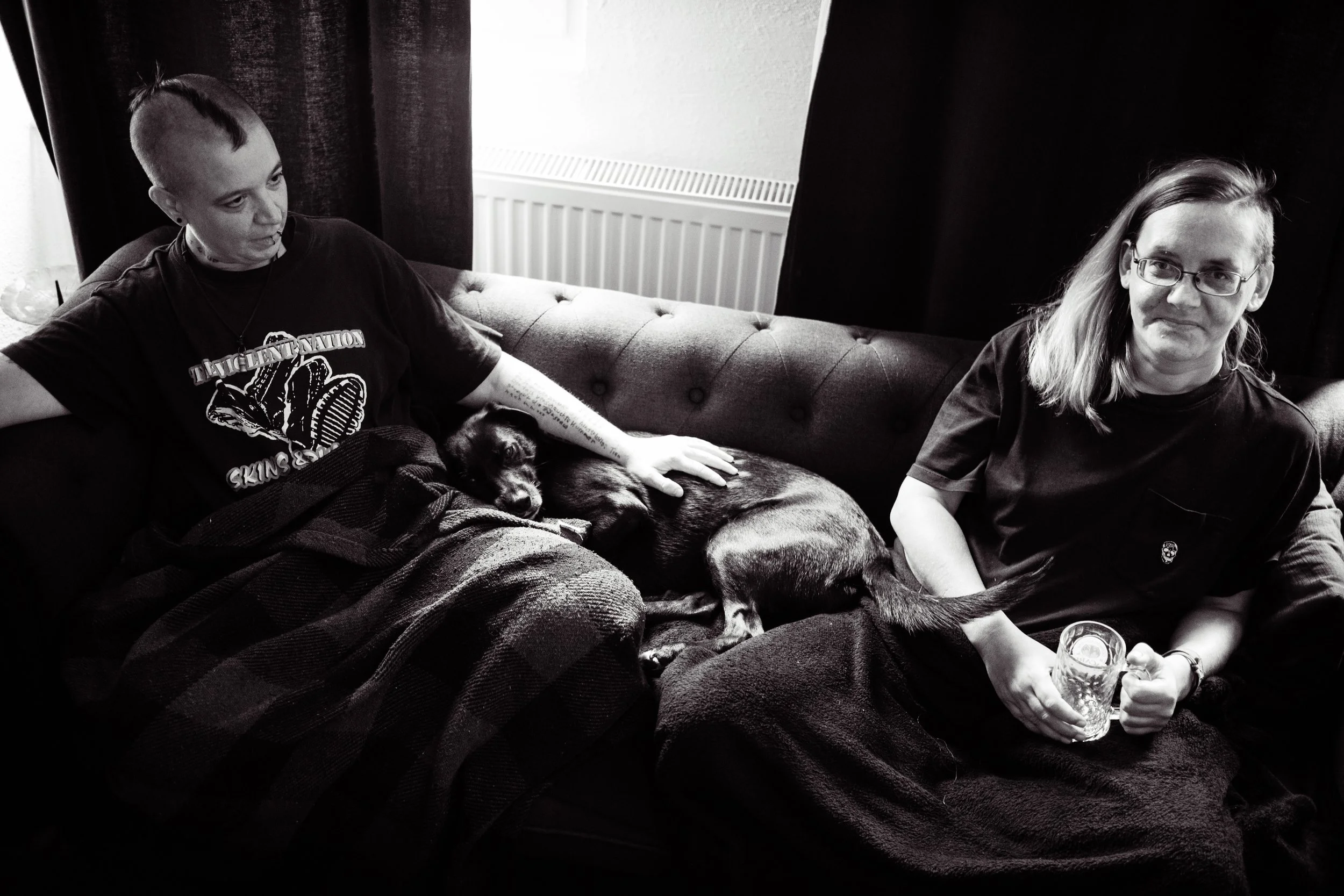 Zwei Menschen sitzen auf einem Sofa, eines hält einen Hund und ein Kind liegt mit dem Hund. Beide haben dunkle Kleidung, im Hintergrund dunkle Vorhänge, der Raum ist hell erleuchtet.